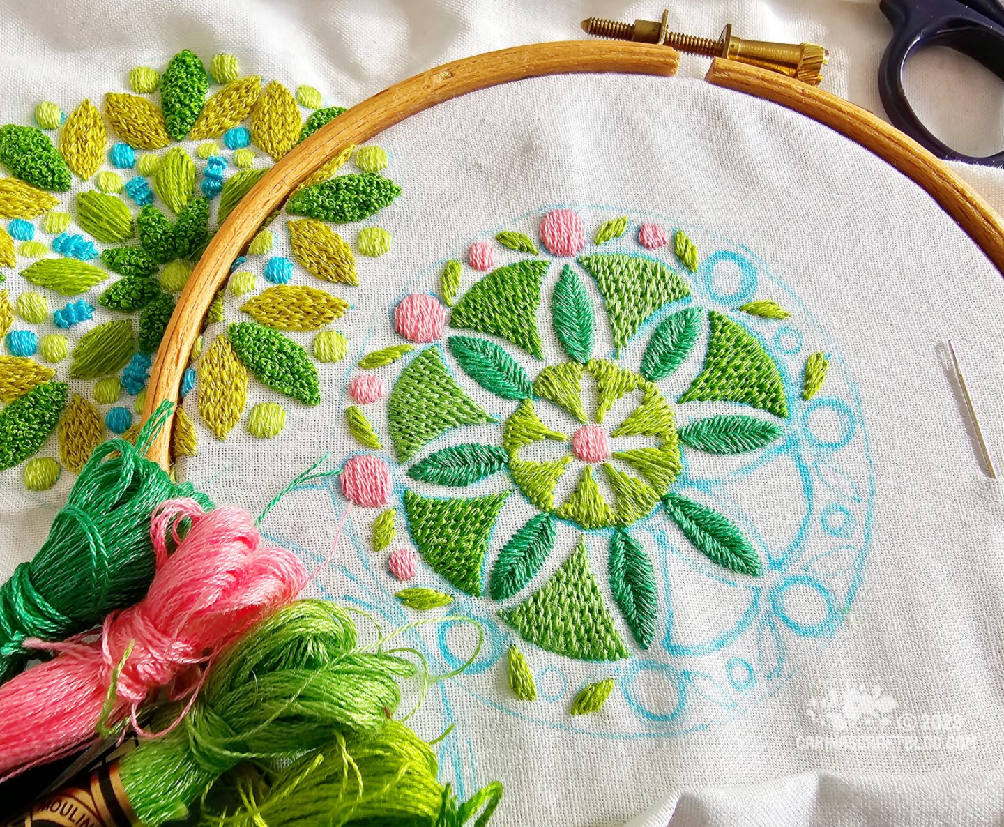 Close view of an embroidery hoop with white fabric. In the centre of the hoop is a partially stitched mandala inspired design in green and pink.