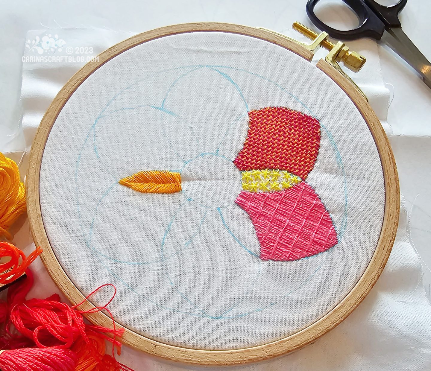 A wooden embroidery hoop with white fabric. On the fabric is a partially embroidered flower shaped design in warm colours using  different stitches.