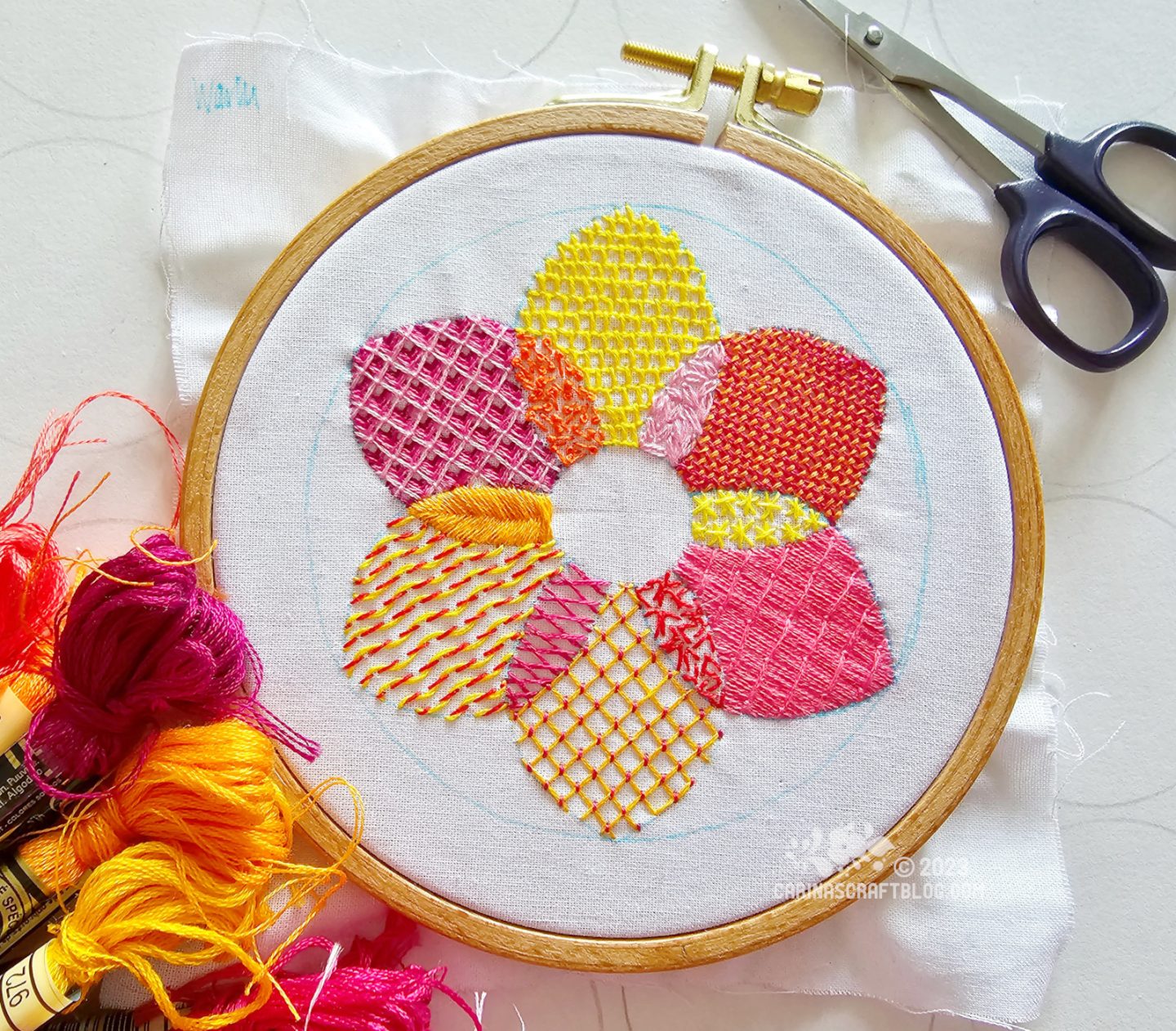 A wooden embroidery hoop with white fabric. On the fabric is embroidered a flower shaped design in warm colours using 12 different stitches.