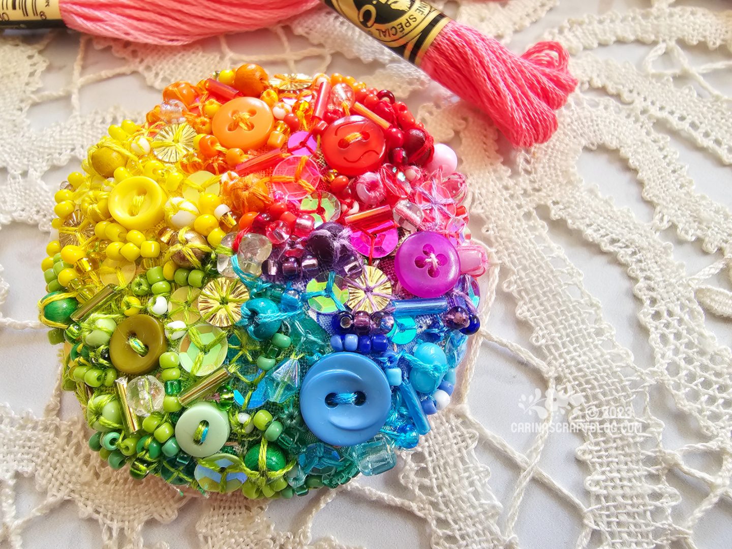 Close view of a circular brooch with a surface made of tiny bits of fabric in a rainbow of colours. The fabric is also decorated with beads and sequins in matching colours.