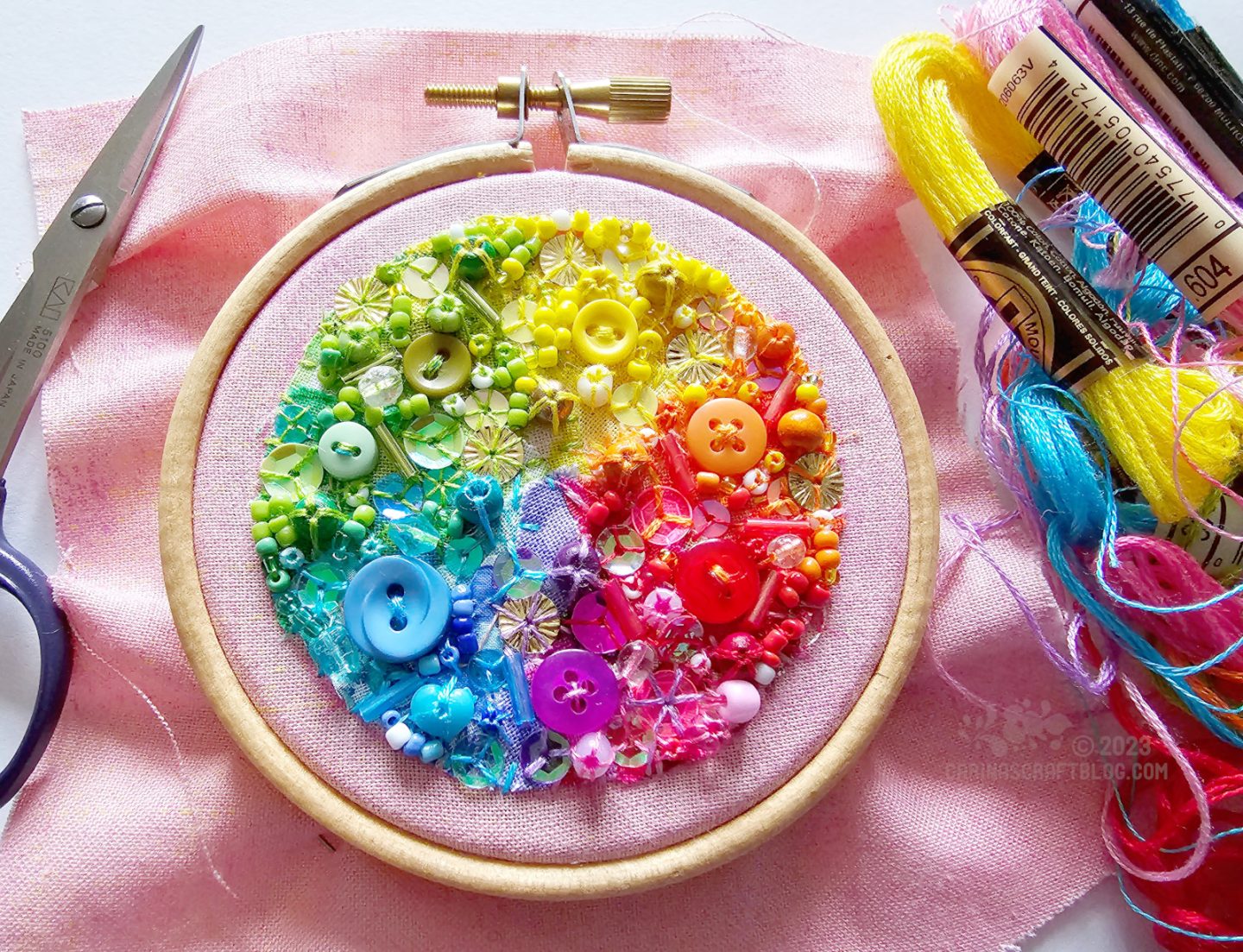 View from above of a small wooden embroidery hoop with light pink fabric. On the fabric is a circle made of tiny bits of fabric in a rainbow of colours. The fabric is also decorated with beads and sequins in matching colours.