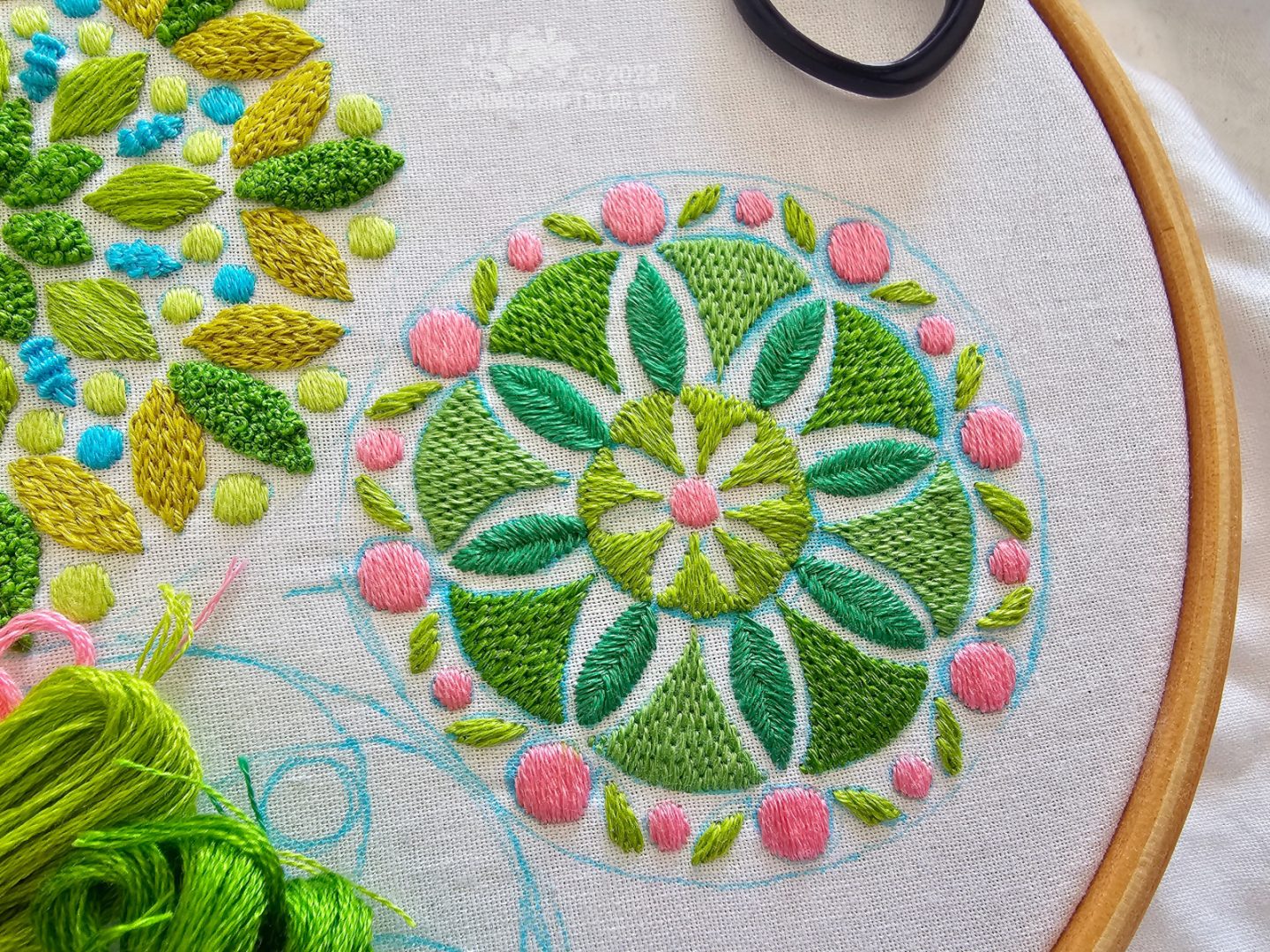 Close view of a mandala inspired design in green colours with pops of pink., embroidered on white fabric.