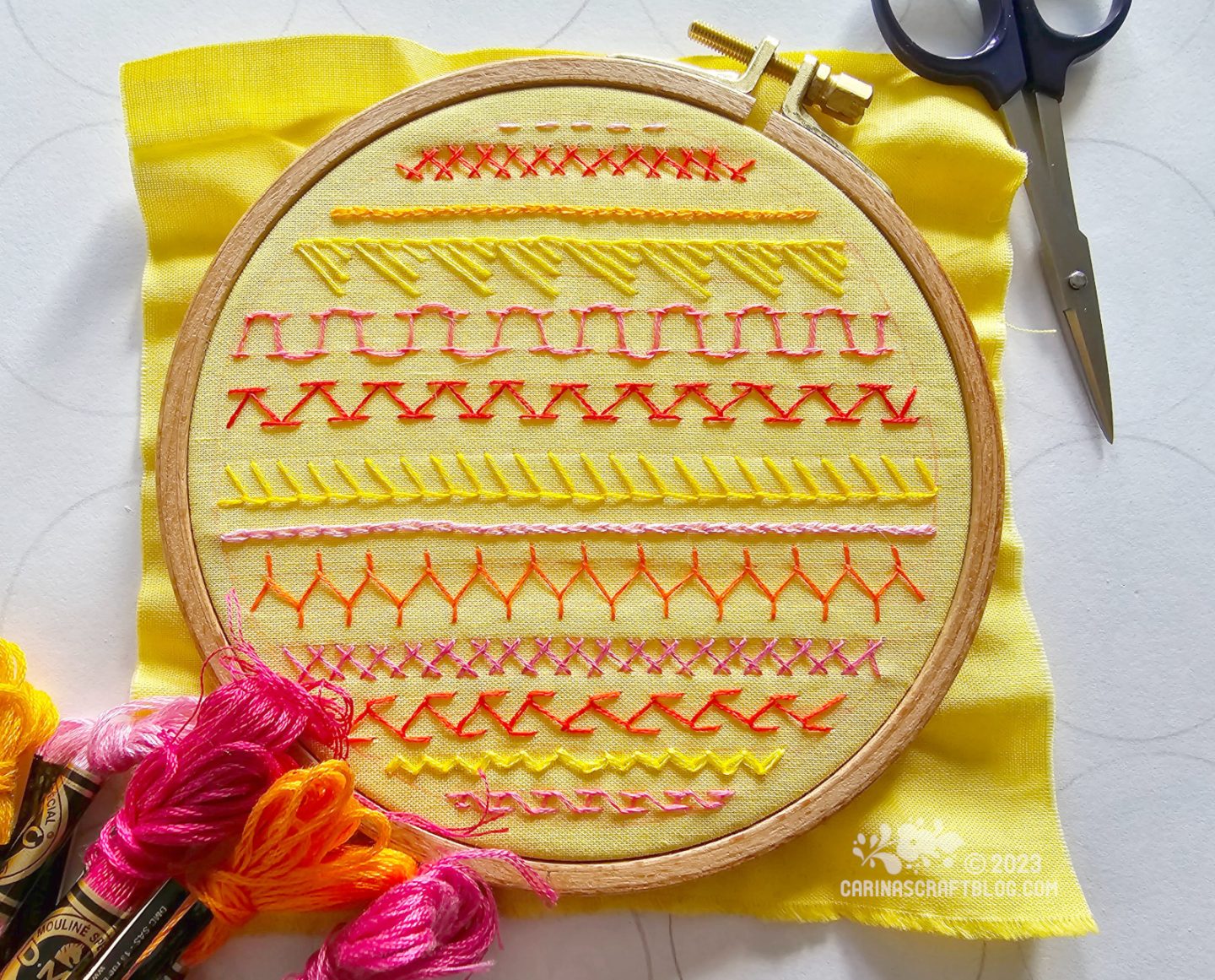 A wooden embroidery hoop with a yellow square of fabric framed in it. On the fabric are stitched rows of different stitches in yellow, orange and pink colours.