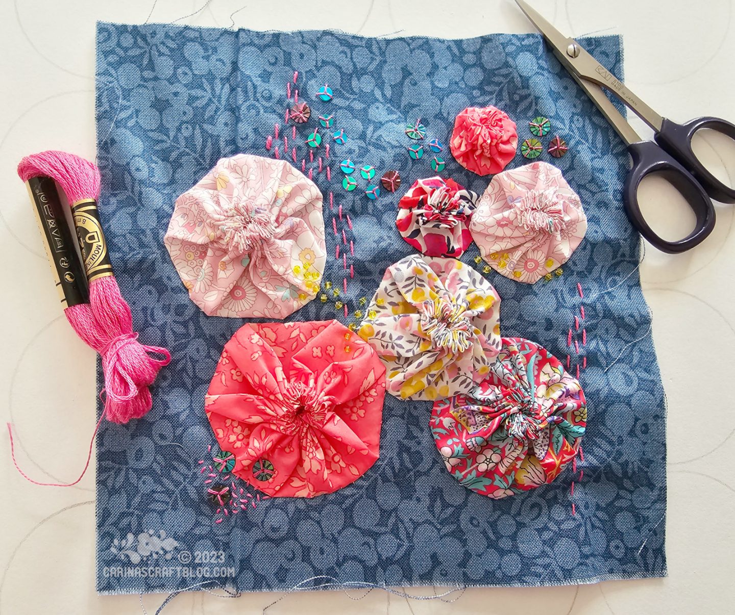 Dark denim blue fabric square decorated with running stitches, sequins and Suffolk puffs in various sizes and pink colours.