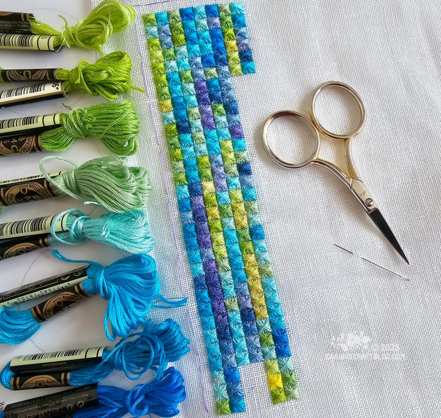 On off white fabric are embroidered columns of square embroidery stitches in various green, blue and purple colours to reflect the daily high and low temperatures. To the left of the stitches are skeins of embroidery thread in some of the same colours.