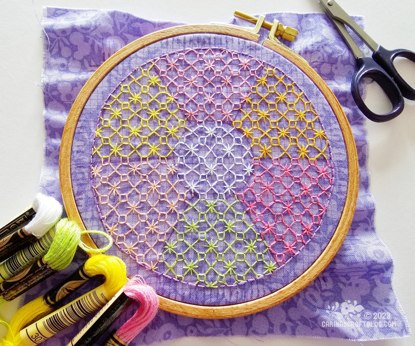 Over head view of wooden embroidery hoop with purple fabric. On the fabric is an embroidered circle that almost fills the hoop. The circle is divided into seven sections, stitched with the same geometric pattern but in seven different colours.