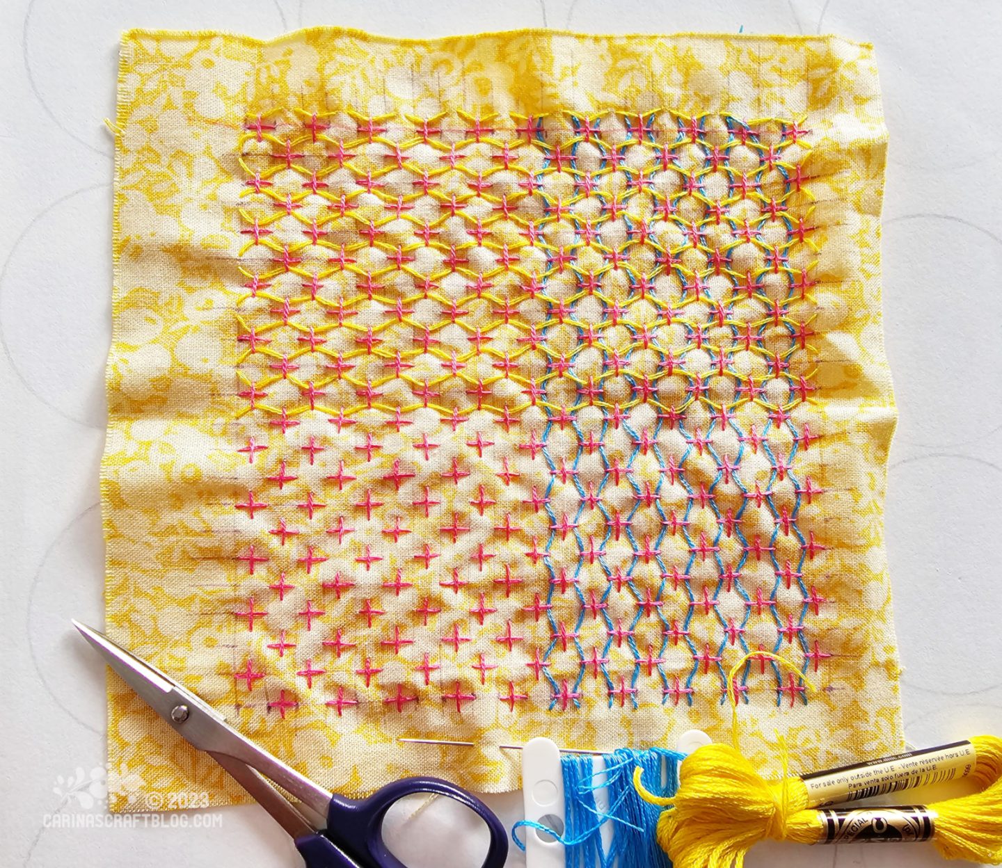 Over head view of a square of yellow fabric. On the fabric is stitched a pattern of crosses with thread woven through the points of the crosses.