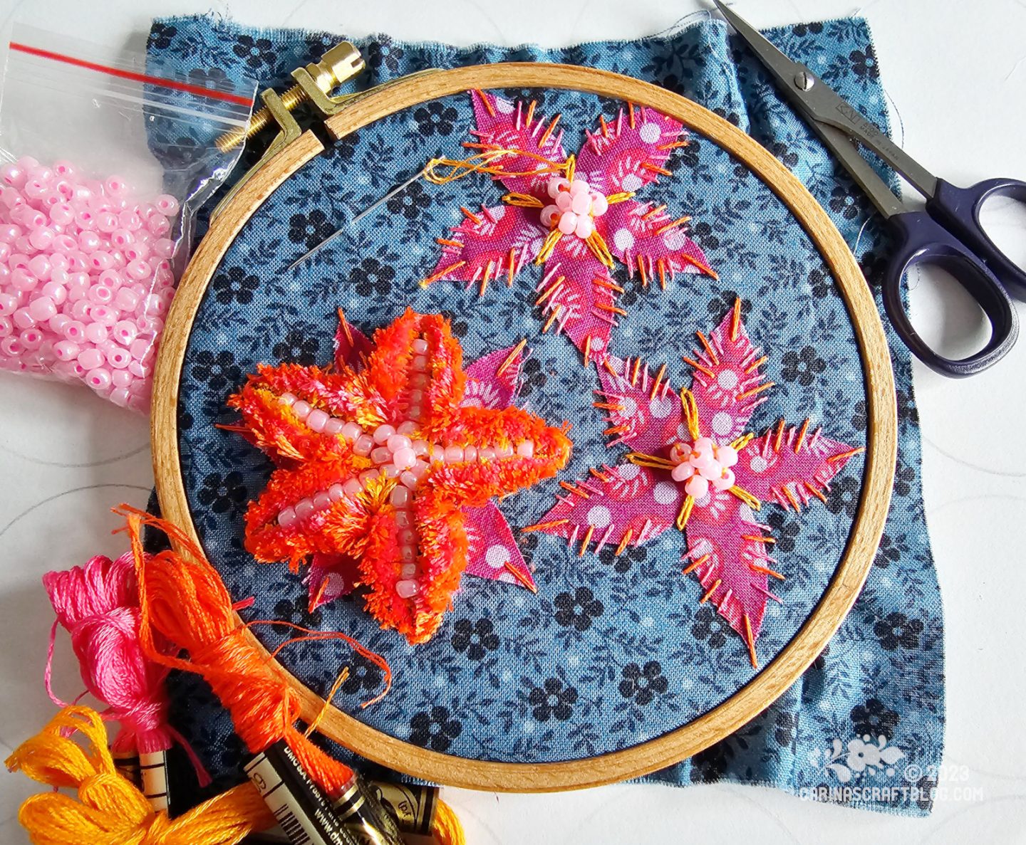 Over head view of wooden embroidery hoop with dark blue fabric. On the fabric are stitched two appliqué flowers with bead decoration. A third flower is worked in tufting stitch which gives it a fluffy appearance, a bit like a fabric pompom.