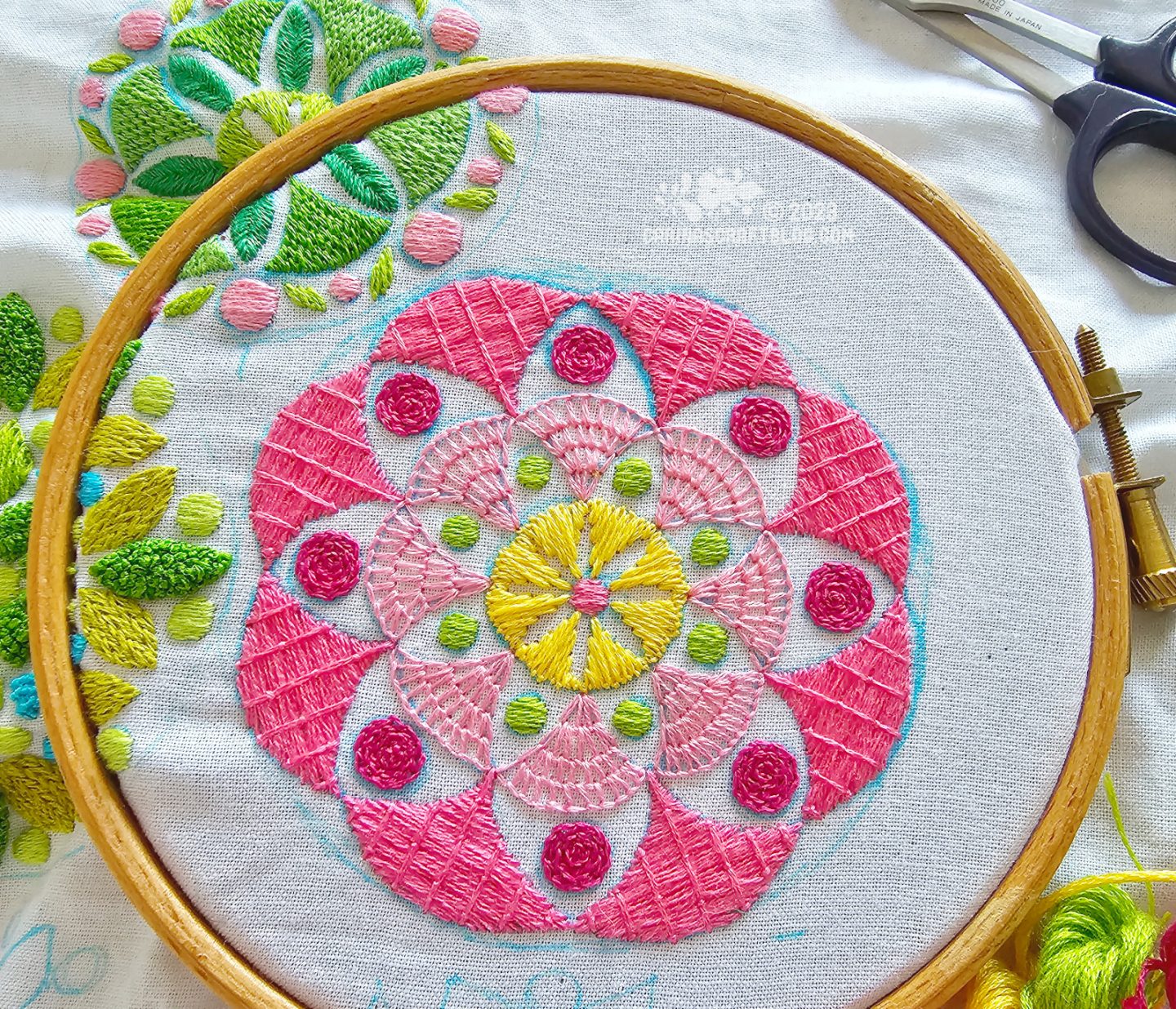 Close overhead view of a wooden embroidery hoop with white fabric. On the fabric is embroidered a mandala inspired design in pink, yellow and green colours.