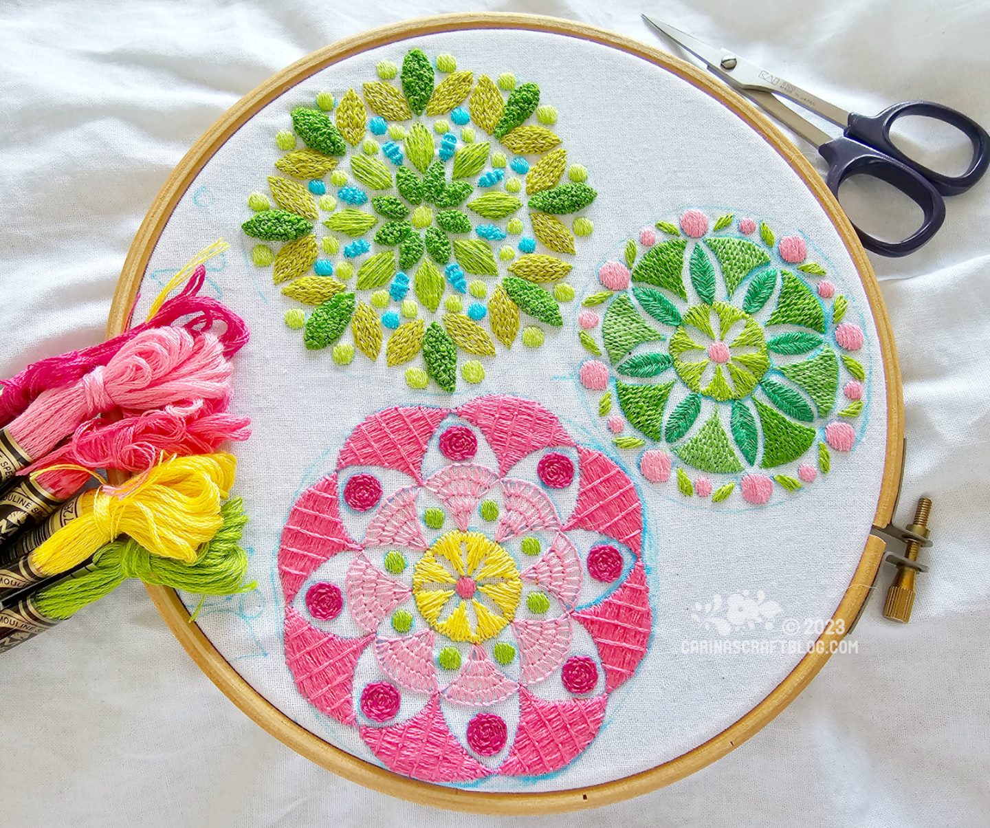 Overhead view of a wooden embroidery hoop with white fabric. On the fabric is embroidered three mandala inspired designs, one mainly in pink colours, the other two in green colours.
