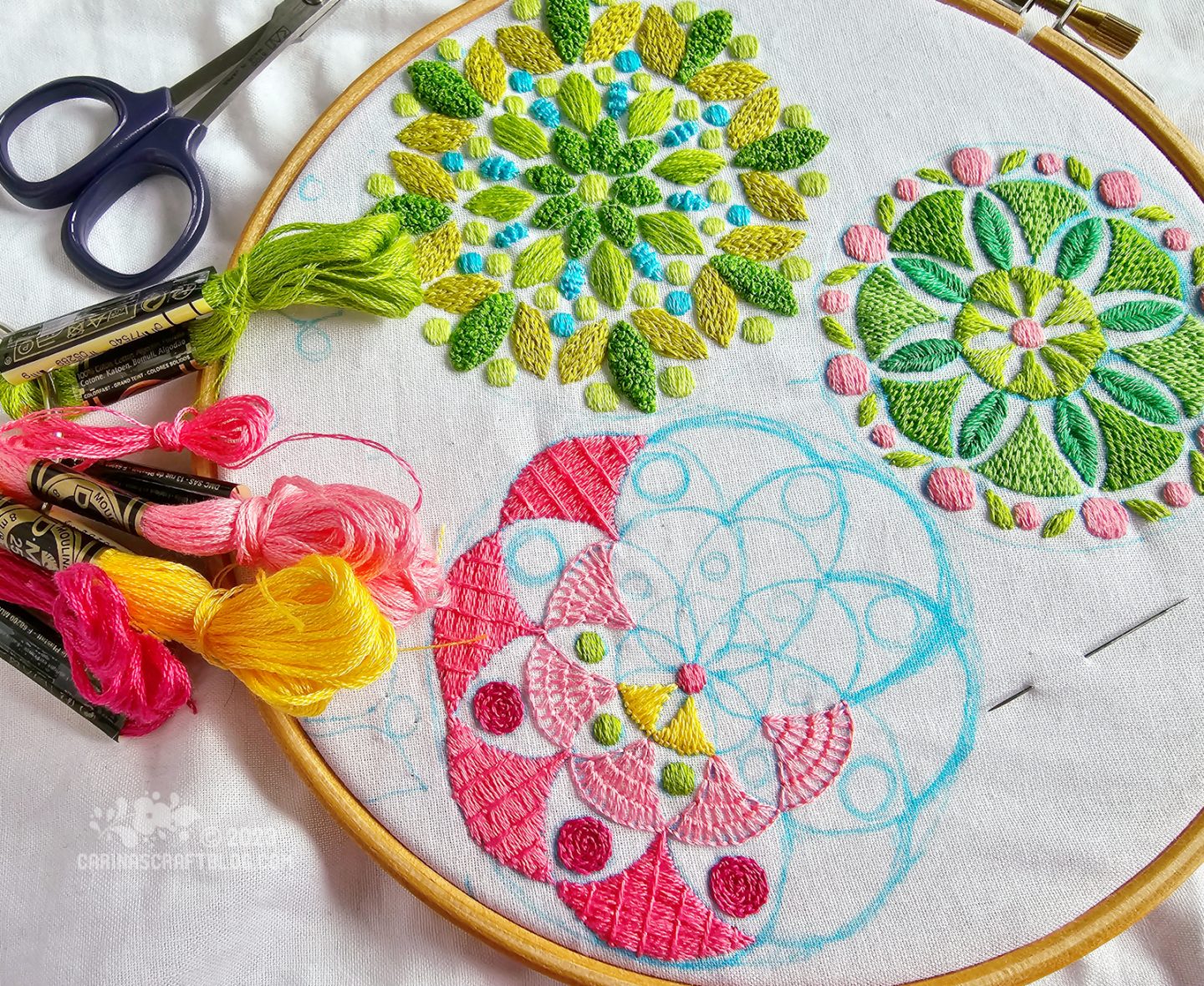 Close view from above of a wooden embroidery hoop with white fabric. On the fabric are embroidered two mostly green mandala inspired round motifs. A third motif is partially embroidered, mainly in shades of pink with accents of yellow and lime green.