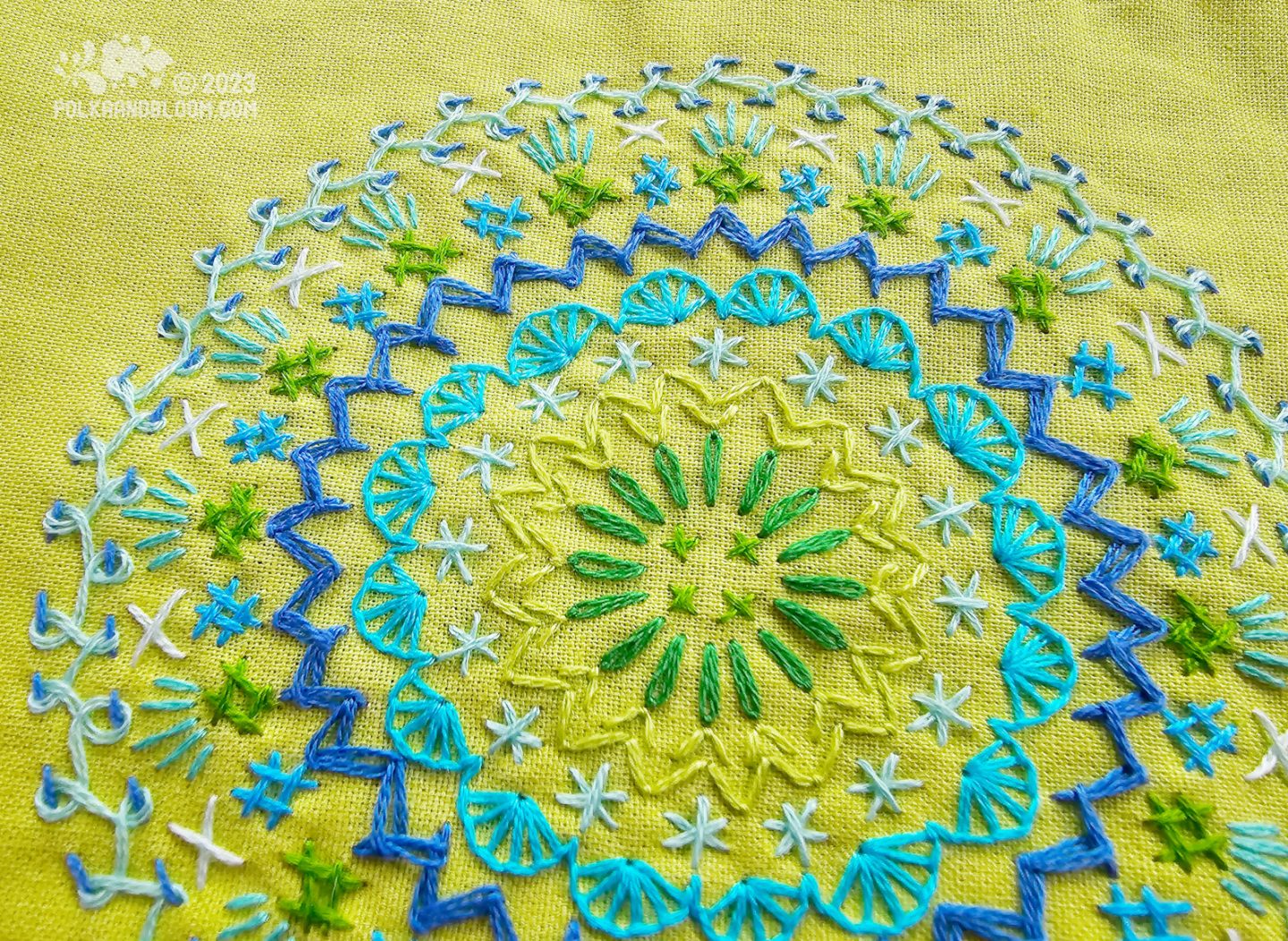 Detail view of a mandala inspired embroidery worked in blue and green colours on a lime fabric.