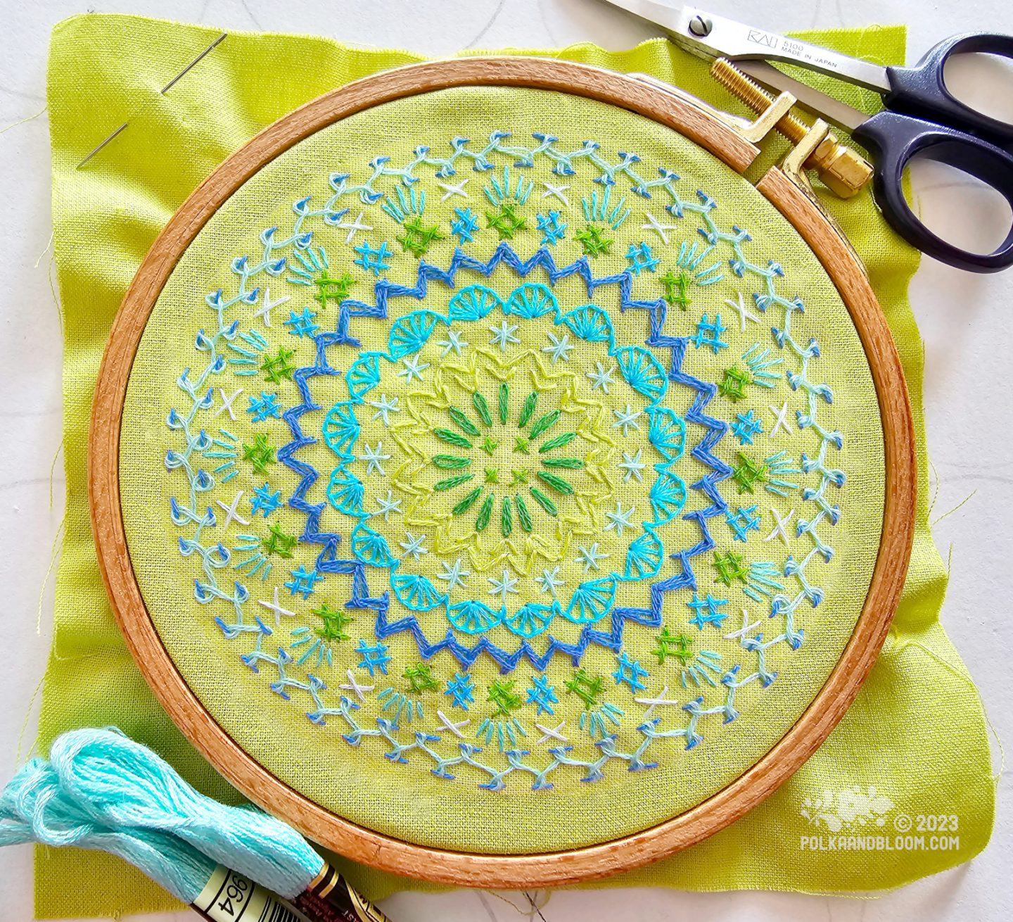 Overhead view of a mandala inspired embroidery worked in blue and green colours on lime green fabric.