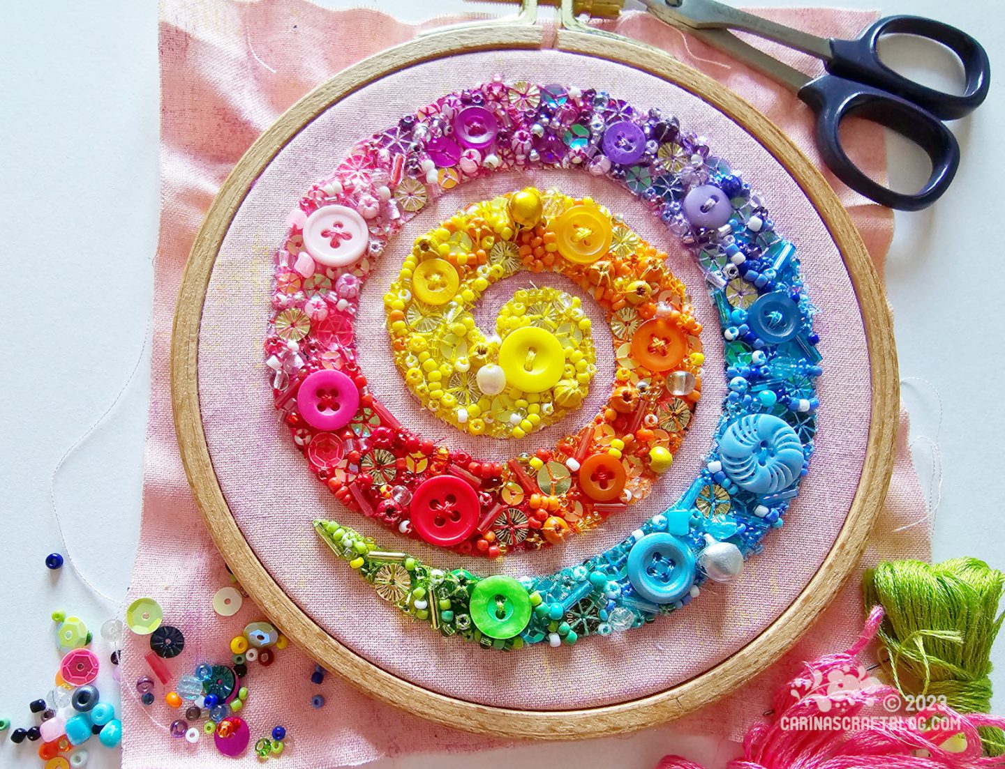 Over head view of wooden embroidery hoop with pale pink fabric. On the fabric is a large spiral filled with fabric, beads and sequins in rainbow order, starting with yellow in the middle and green at the outer end.