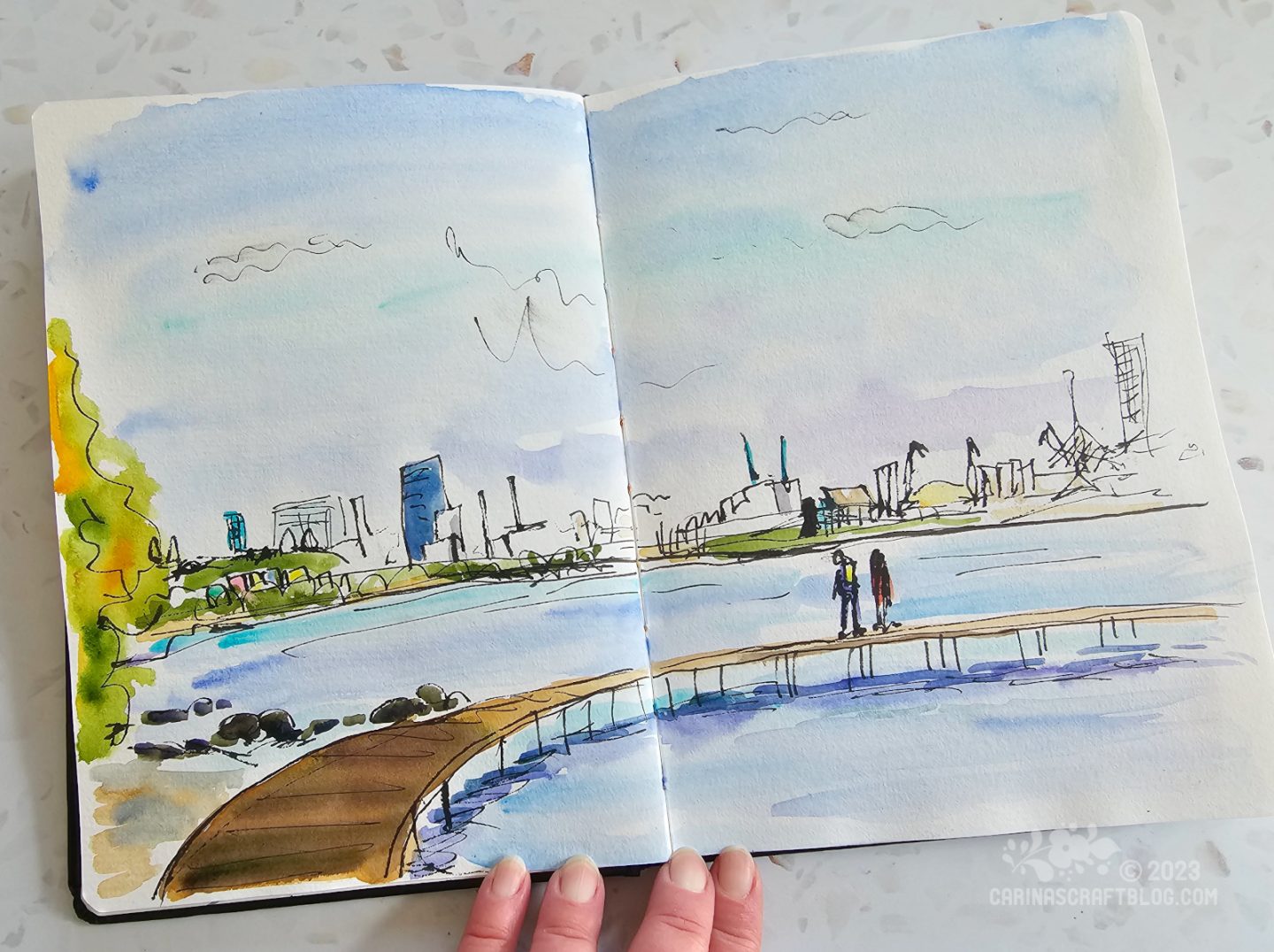 Photo of an open spread in a sketchbook. The spread shows a sketch of a beach with a wooden bridge in the foreground and the skyline of a city in the background.