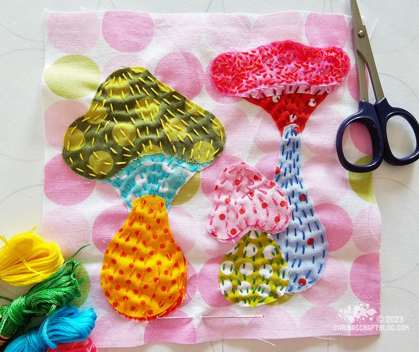 Over head view of a square piece of fabric with a print of pink circles. On the fabric are appliquéd simplified mushrooms using bright fabrics all printed with dots in different sizes.