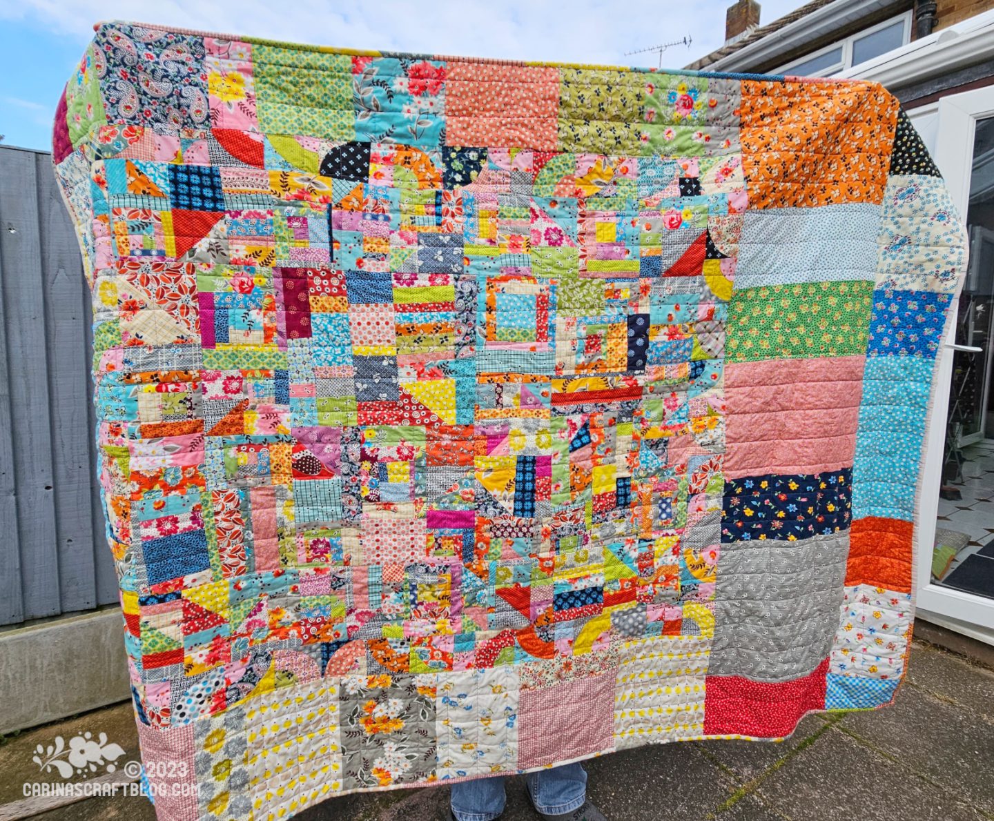 A quilt is held up horizontally by a person standing on a patio with a blue fence behind them. The quilt back is made up of multi colour blocks in various shapes and sizes.