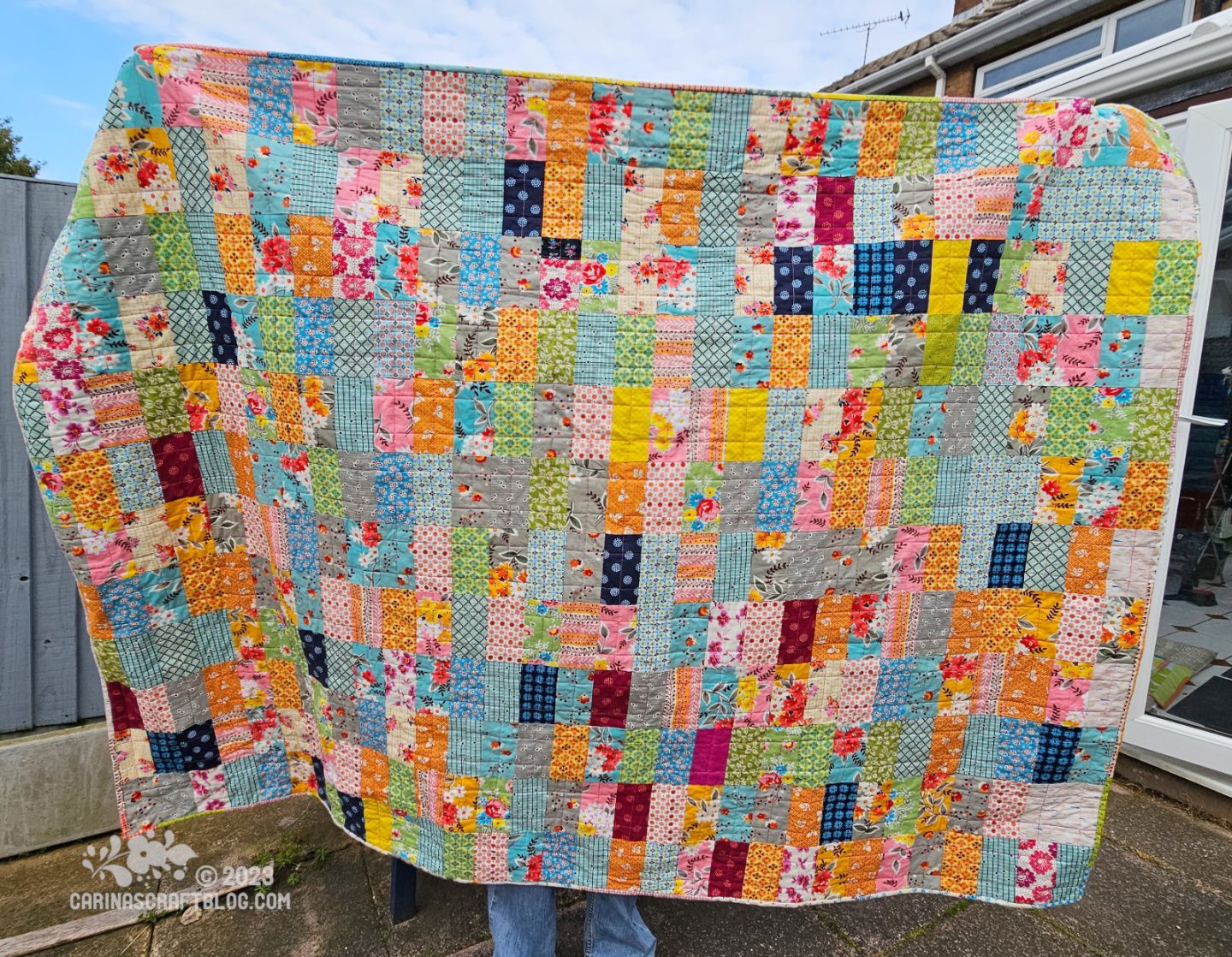 A quilt is held up horizontally by a person standing on a patio with a blue fence behind them. The quilt front is made up of multi colour rectangles. 