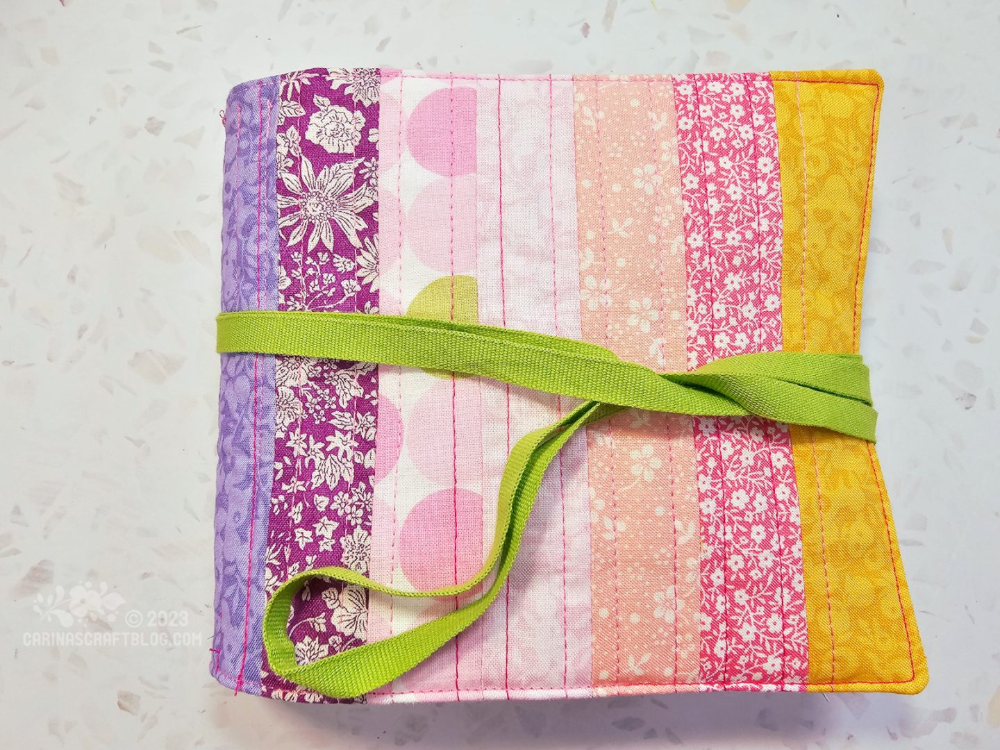 Over head view of a textile book. The cover of the book is made of vertical strips of fabric in purple, pink and yellow colours. A narrow lime green ribbon is wrapped around the book.
