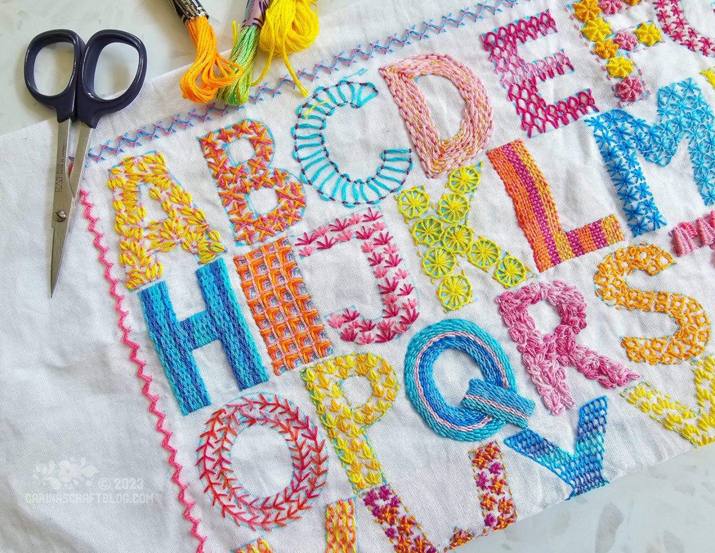 Overhead, partial view of an alphabet design with block letters embroidered with pink, yellow, blue and orange colours on white fabric.