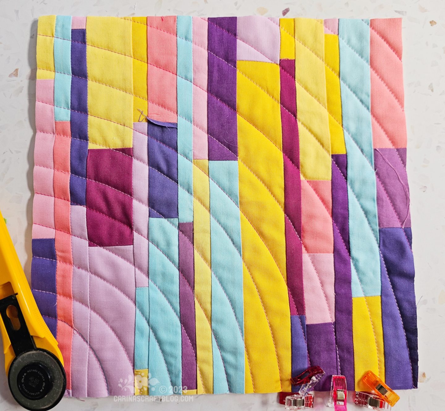 Small square quilt of narrow vertical strips in blue, pink, purple and yellow colours.