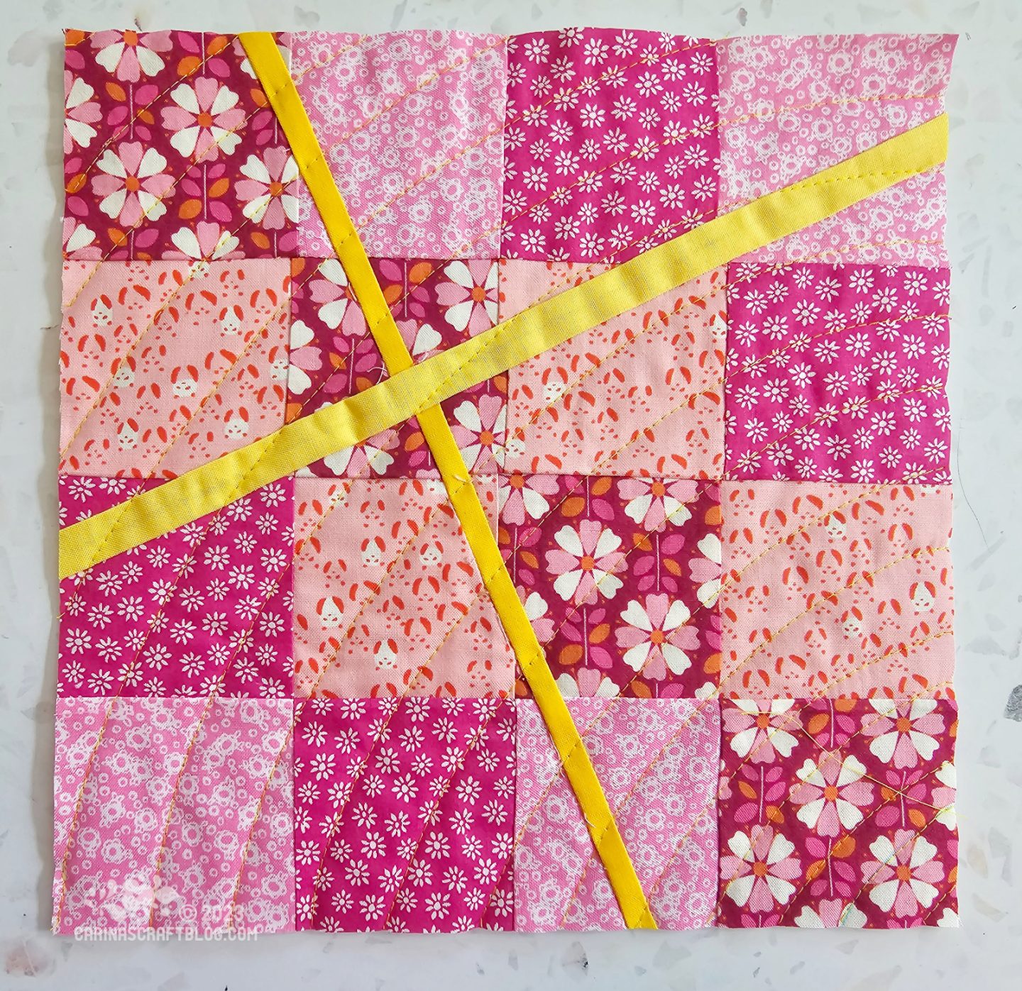 Small square quilt made of 16 pink squares of fabric in a four by four grid. The grid has been slashed across in two random diagonals and two yellow strips have been inserted in the grid.