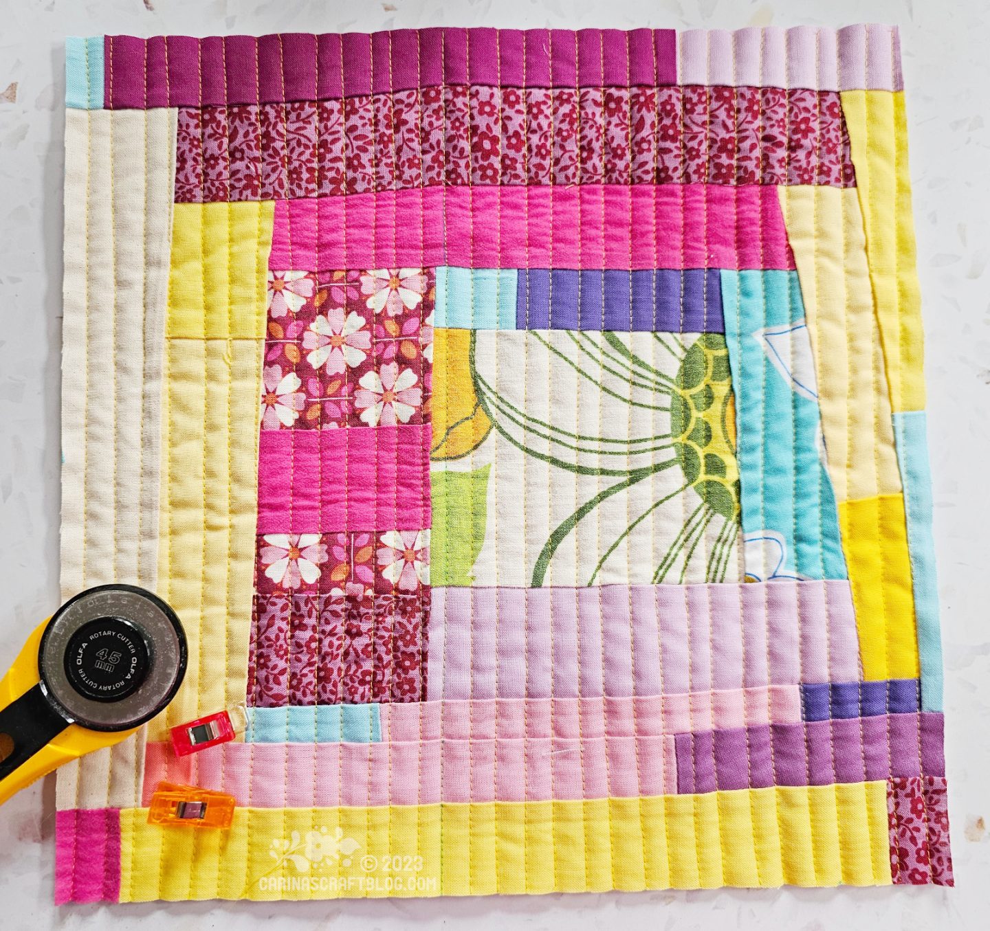 Small square quilt with a small white square in the centre, surrounded by strips of different thicknesses in mainly yellow, pink, purple and maroon colours.