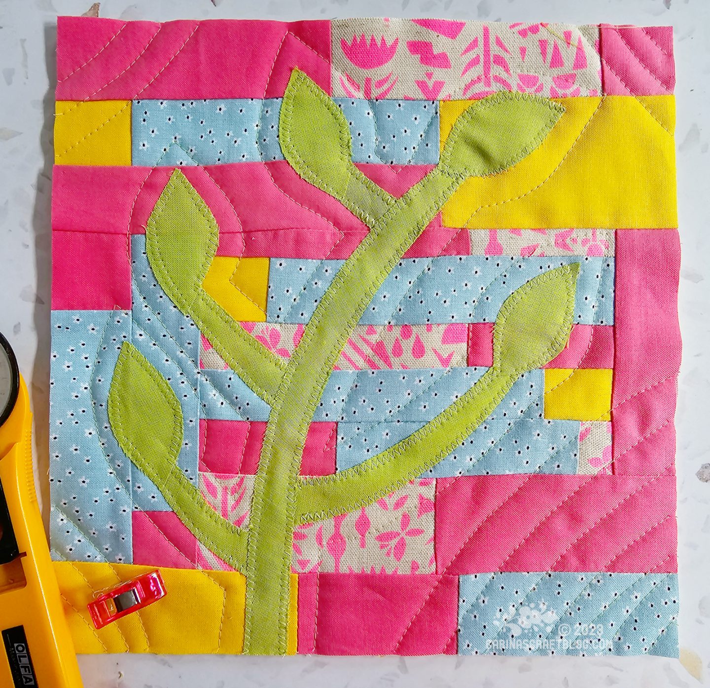 Small square quilt made of narrow strips in pink, blue and yellow, with a green branch appliquéd on top.