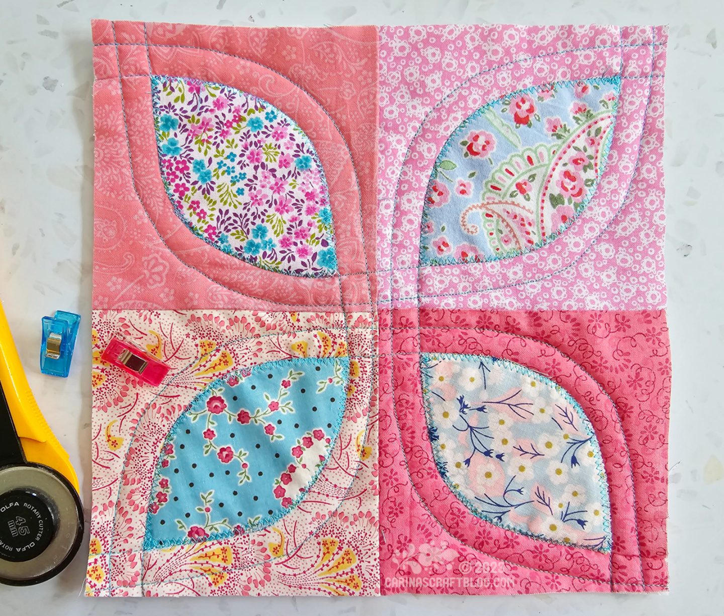 A small square quilt made of four different squares of pink with a blue leaf shape appliquéd in each square.