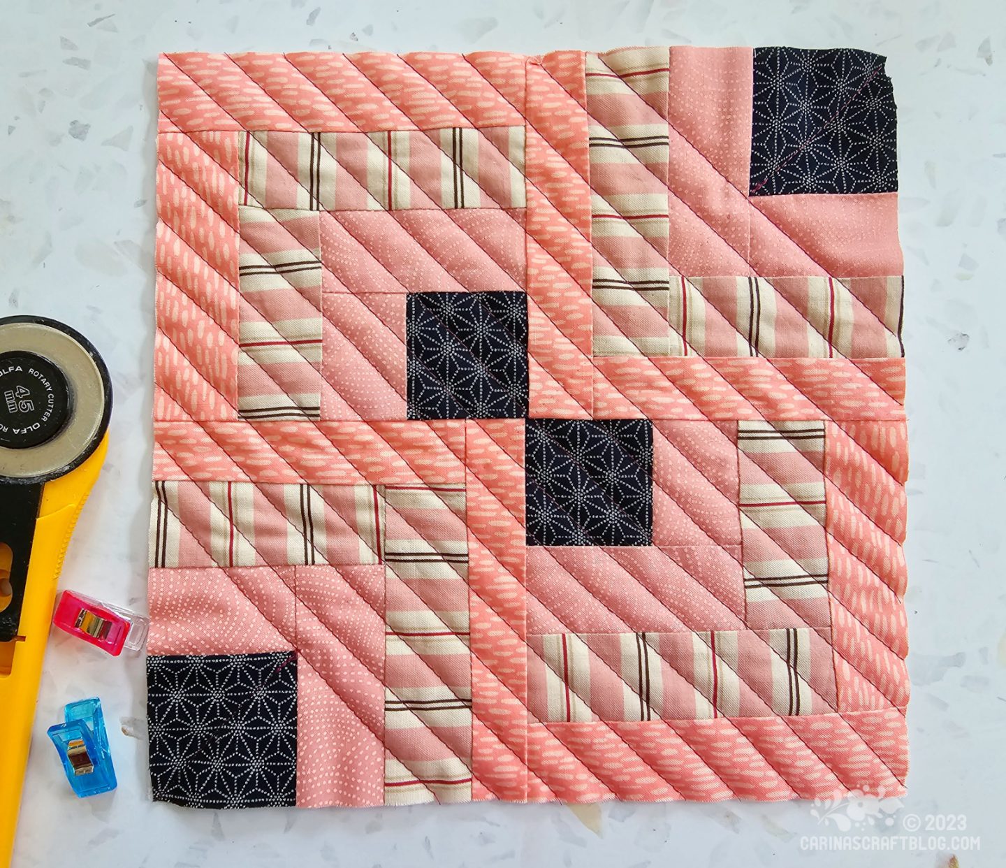 A small square quilt made of four square blocks each with a dark blue square in one corner, surrounded by strips of salmon coloured fabrics.