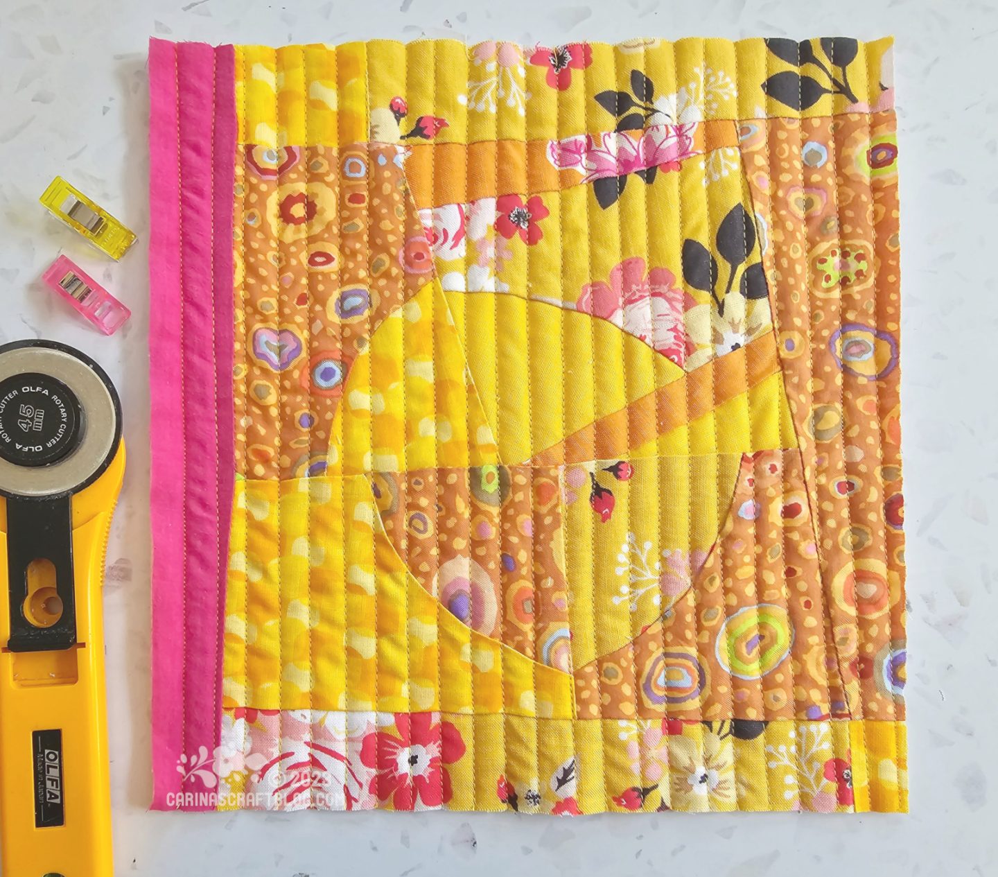 Small square quilt made with yellow fabrics, making a wonky multi part circle in the centre, surrounded by strips and triangles to make the square. On the left is a vertical solid hot pink strip.