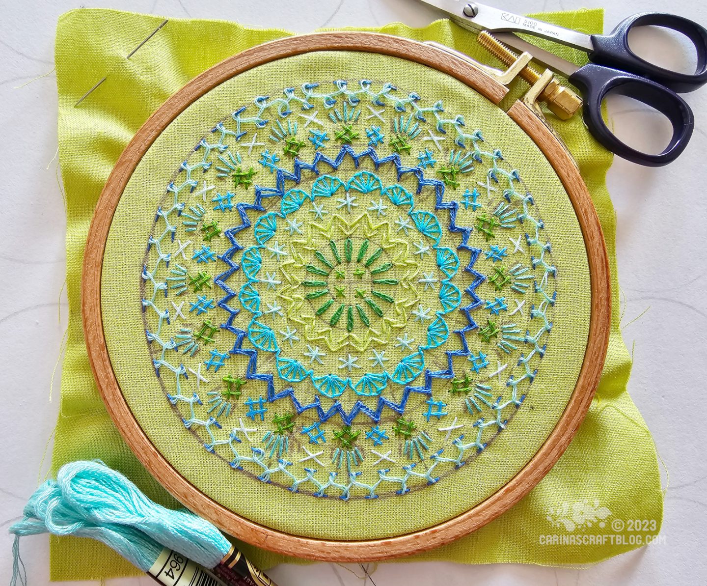 Overhead view of a wooden embroidery hoop with lime green fabric. On the fabric is embroidered a mandala inspired design in blue and green colours.