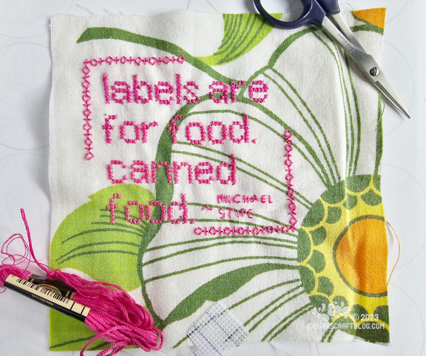Close overhead view of a square of fabric showing a partial print of a large flower. On the fabric is a quote stitched in cross stitches: Labels are for food, canned food. Michael Stipe.