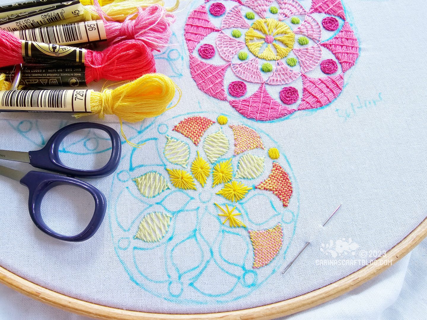 Overhead, partial view of a large embroidery hoop with white fabric. On the fabric are embroidered two mandala inspired designs, one in mainly pink colours, one in mainly yellow colours.