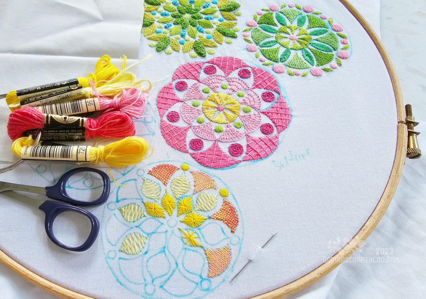 Overhead, partial view of a large embroidery hoop with white fabric. On the fabric are embroidered four mandala inspired designs, two in green colours, one in mainly pink colours and one in mainly yellow colours.