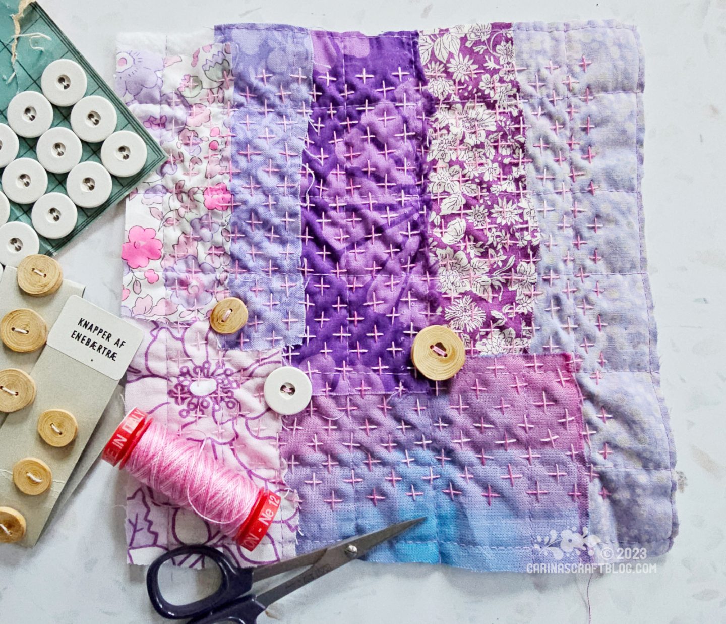 Overhead view of a small wonky, raw edge patchwork made of several different fabrics in purple colours. The patchwork is quilted with cross shaped stitches and decorated with three buttons.