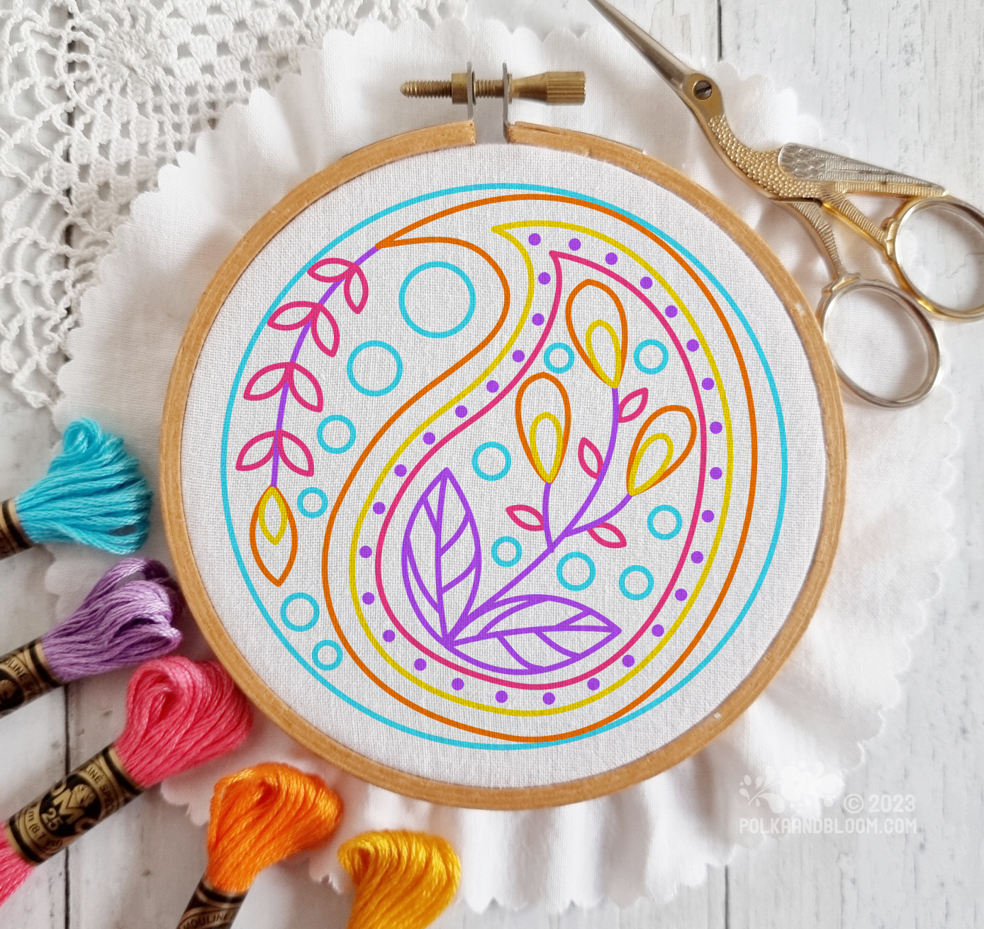 Small wooden embroidery hoop seen from above. There is white fabric in the hoop and the image is overlaid with a line drawing of a paisley design in blue, yellow and purple.