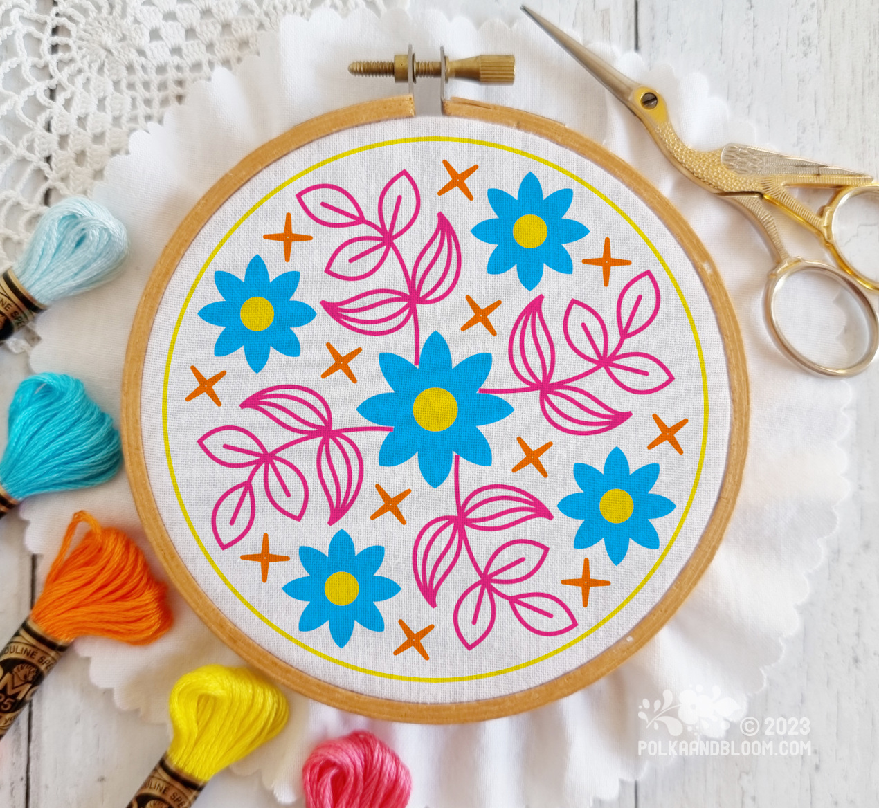 Small wooden embroidery hoop seen from above. There is white fabric in the hoop and the image is overlaid with a line drawing of blue flowers with pink leaves.