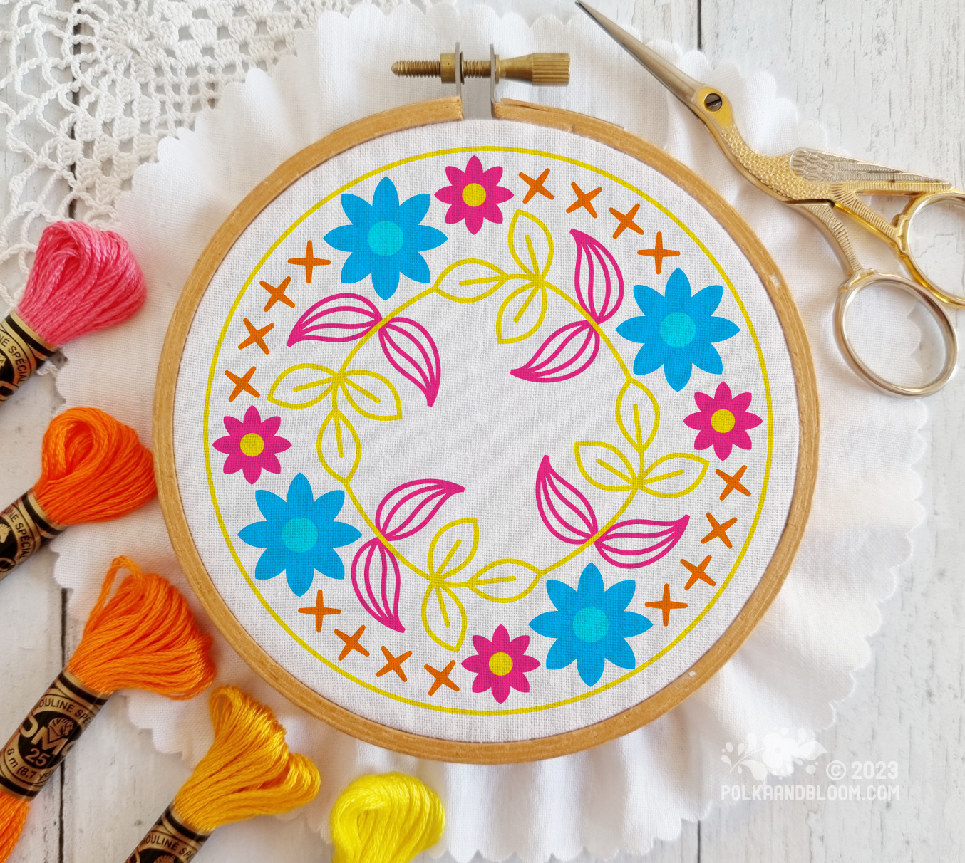 Small wooden embroidery hoop seen from above. There is white fabric in the hoop and the image is overlaid with a line drawing of blue flowers with pink and yellow leaves.