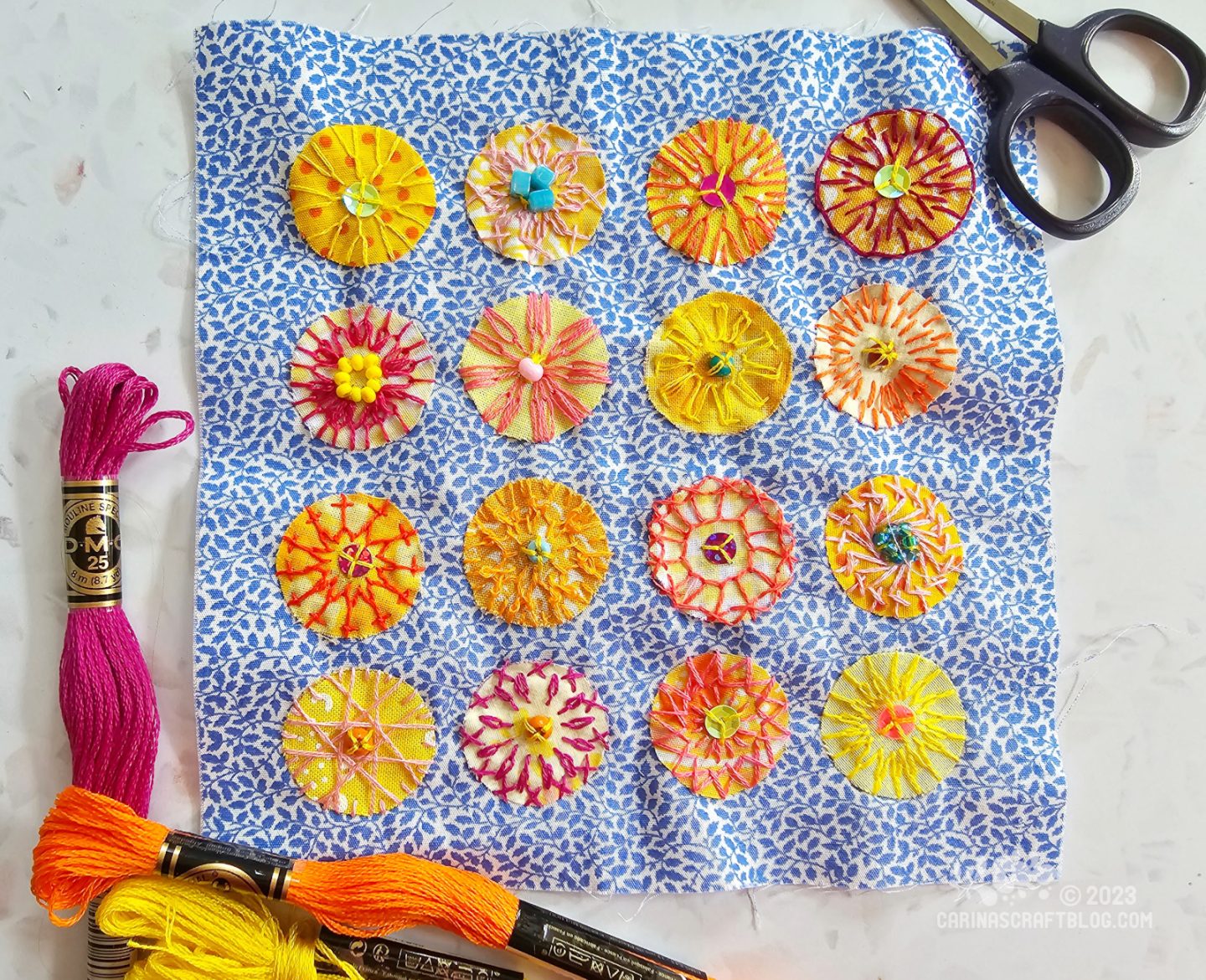 Overhead view of a square of fabric printed with a tiny blue leaf design. 16 yellow or orange fabric circles are appliquéd on the fabric in a 4 by 4 grid. The circles are decorated with various stitches and beads.