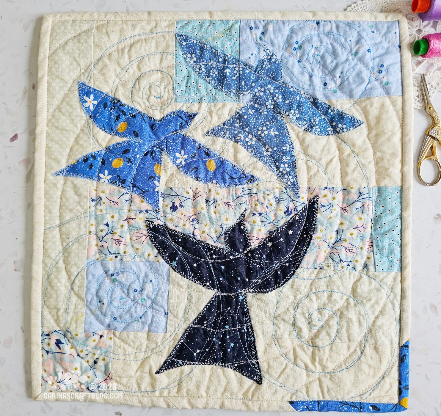 View from above of a mini quilt. The background is a random patchwork in white and light blue with three birds in different shades of blue appliquéd on top.
