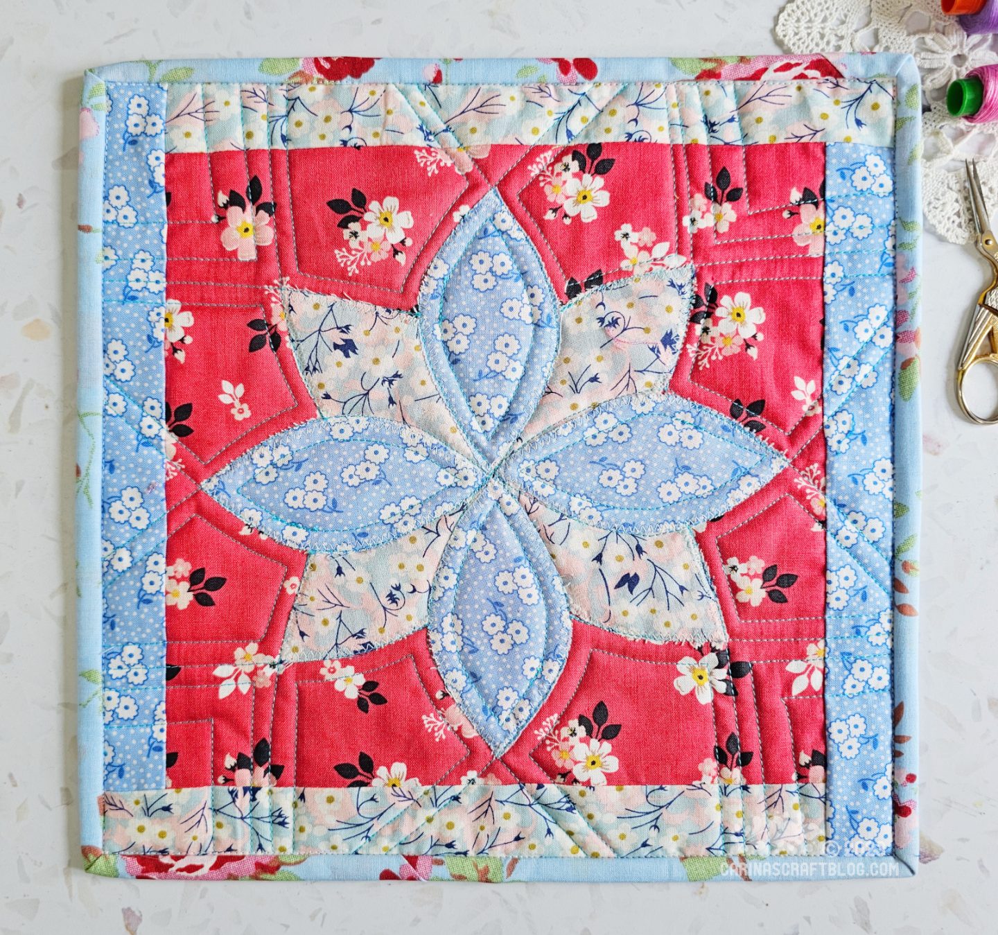 View from above of a mini quilt. The background is red fabric with white and black flowers. On that is appliquéd a sort of flower shape in light blue colours. There is a border in the same light blue fabrics.