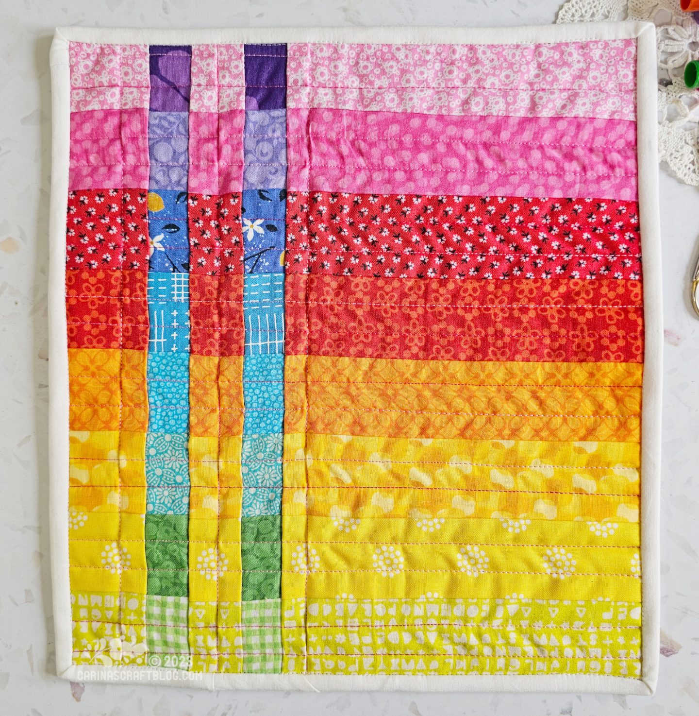 View from above of a mini quilt. quilt is made of horizontal strips of warm colours in rainbow colour order from pink at the top to green at the bottom, inset with two strips of cool colours.