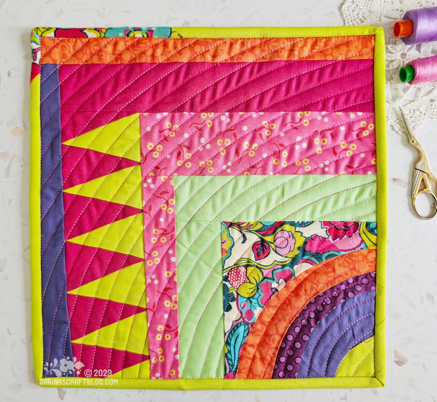View from above of a mini quilt. Starting with wonky concentric quarter circles in the right bottom corner, turning to quarter squares with a section of isosceles triangles. The main colours are hot pink, lime green, orange and purple.