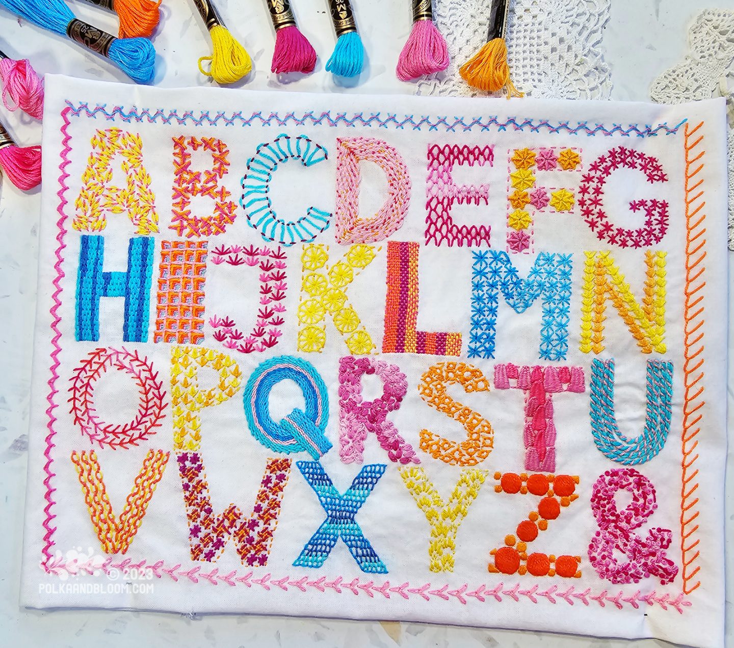 Overhead view of a rectangle of white fabric embroidered with the letters of the English alphabet plus the ampersand.