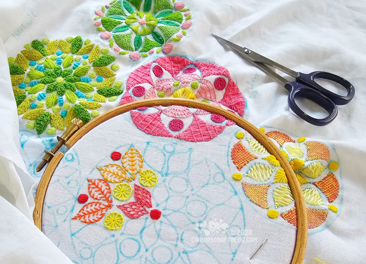 Overhead view of an embroidery hoop with white fabric embroidered with round mandala inspired motifs in pink, yellow and green colours.