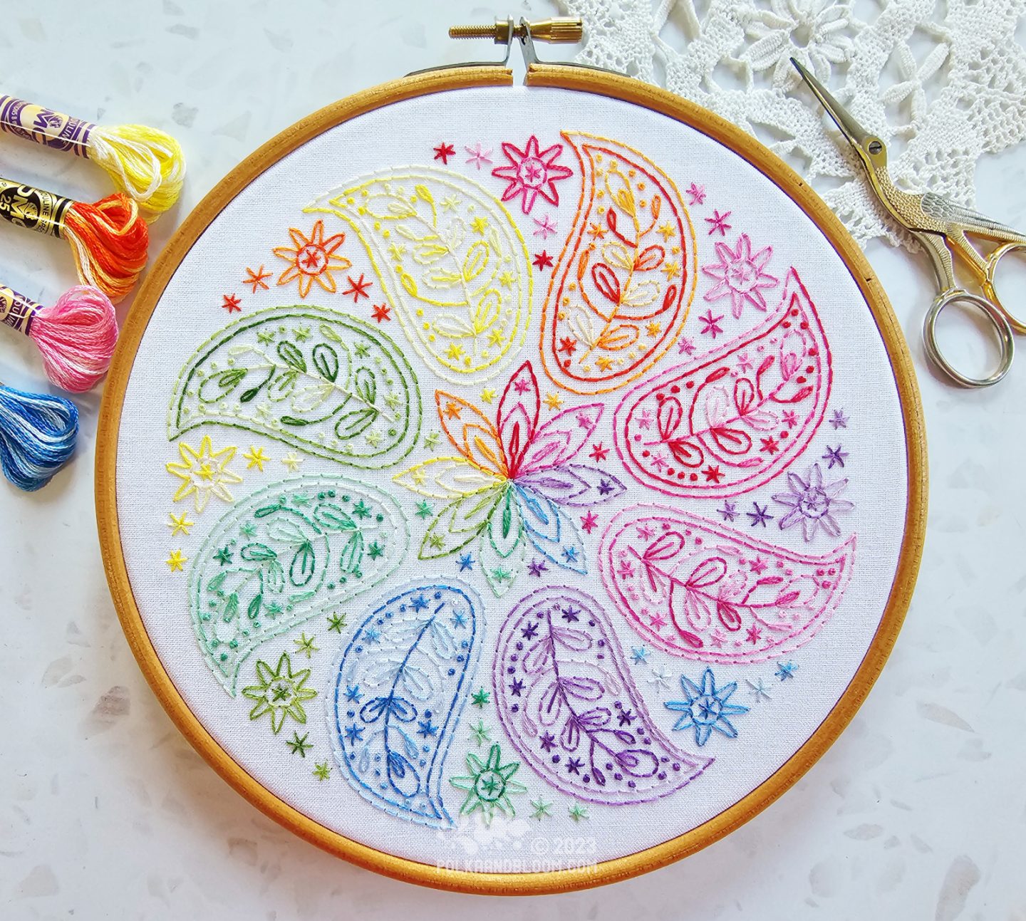 Overhead view of an embroidery hoop with white fabric. On the fabric is embroidered a mandala inspired design with eight small paisley shapes each in a different colours in rainbow order.