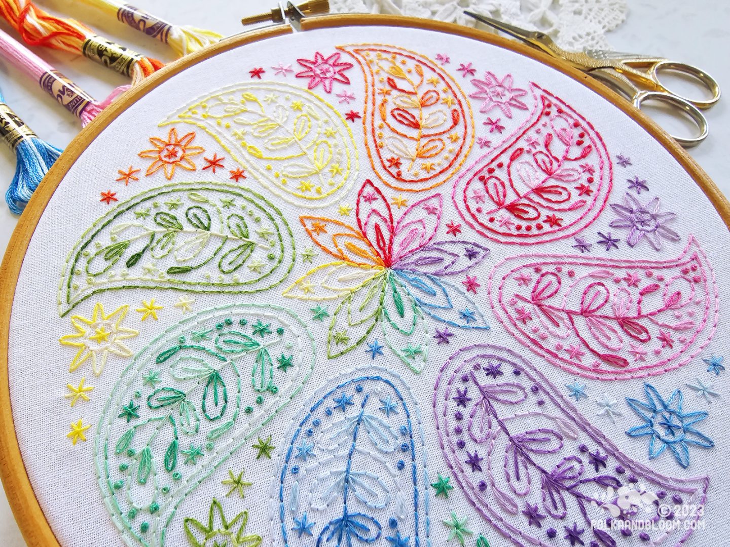 Close up view of an embroidery hoop with white fabric. On the fabric is embroidered a mandala inspired design with eight small paisley shapes each in a different colours in rainbow order.