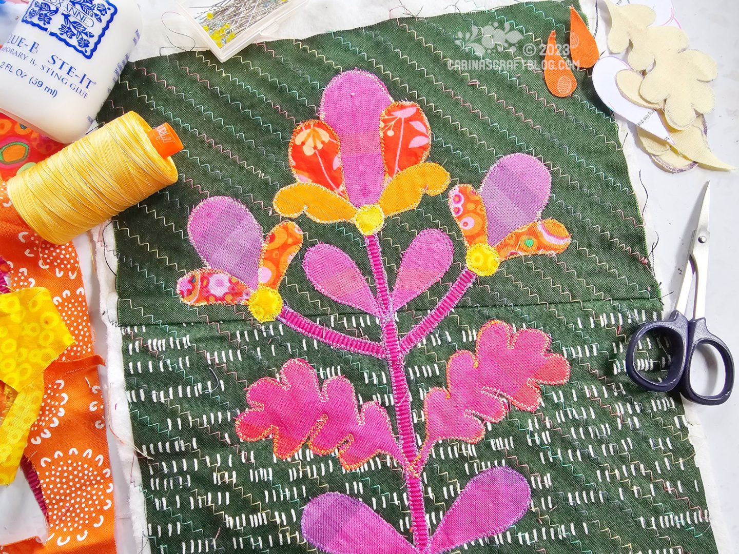 Overhead view of a green piece of fabric quilted with zigzag stitching and appliquéd with a flower in magenta and orange colours.