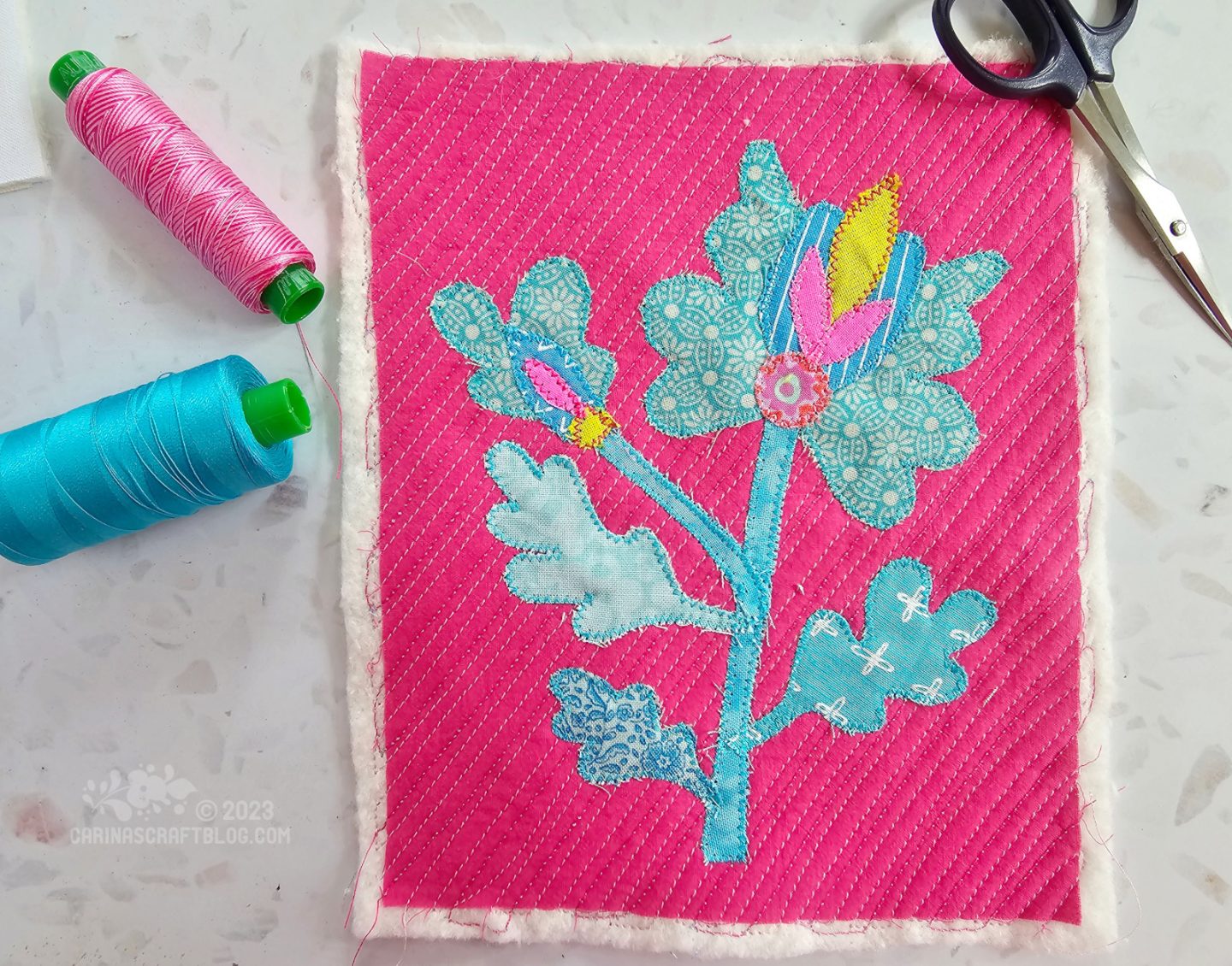 Overhead view of a quilted piece of hot pink fabric with a blue flower appliquéd on top.